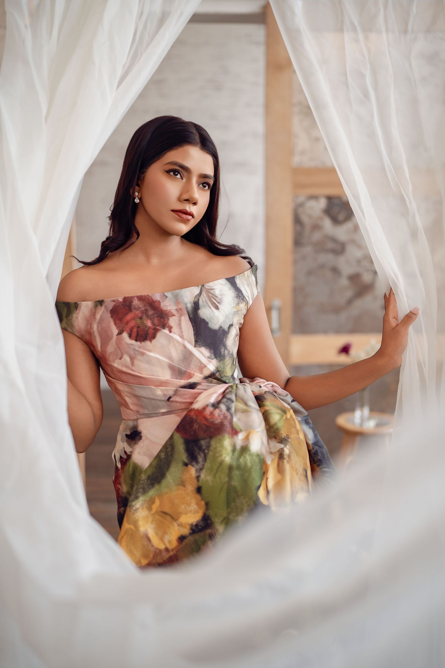 Woman in floral off-shoulder dress poses elegantly between sheer white curtains in a softly lit room.