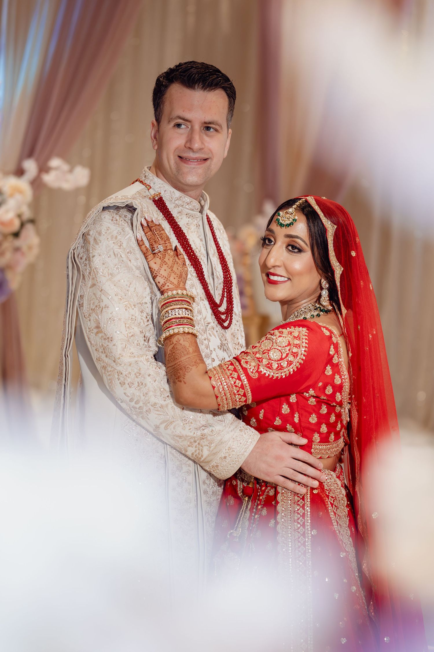 Couple in traditional Indian wedding attire, bride in red lehenga with gold jewelry, groom in cream sherwani, posing together.