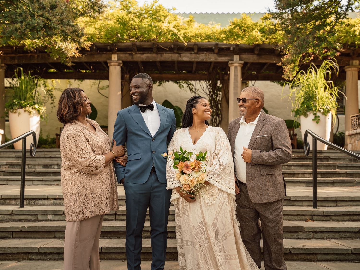 A joyful wedding party with bride, groom, and two guests posing on outdoor steps surrounded by lush greenery.