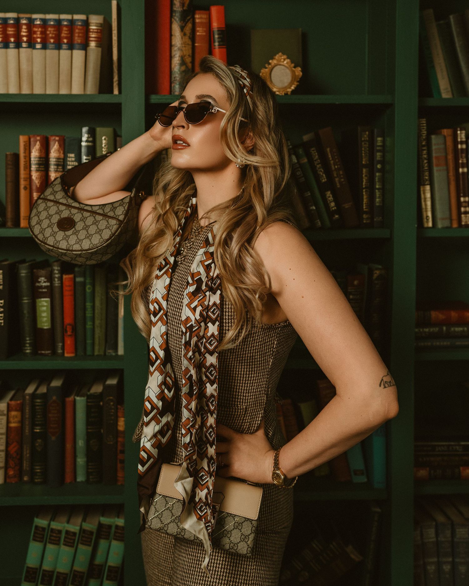 Stylish woman in vintage-inspired outfit with sunglasses posing in front of green bookshelf filled with books.