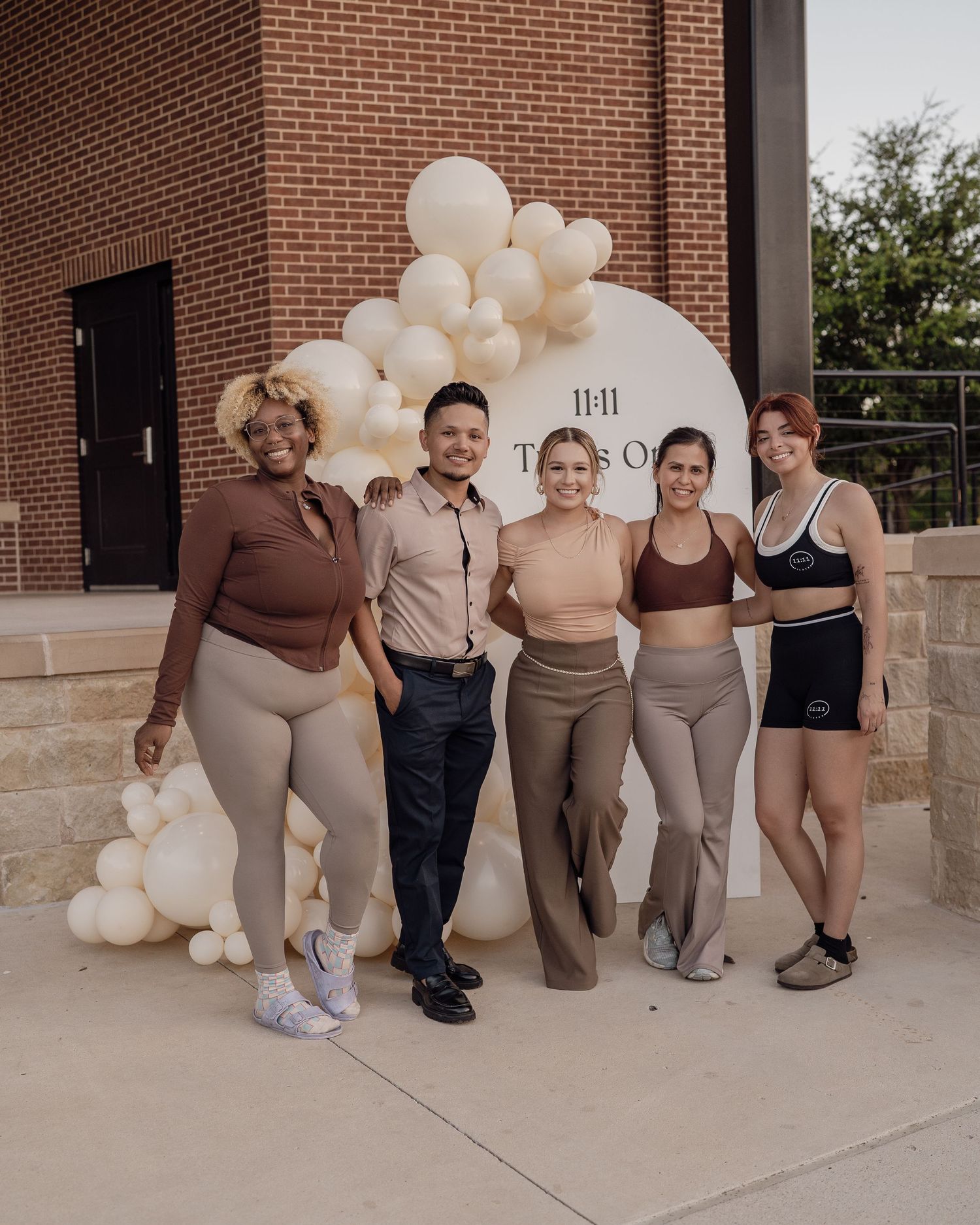 Five people posing together outdoors in front of a balloon arch and circular sign at a fitness studio event.