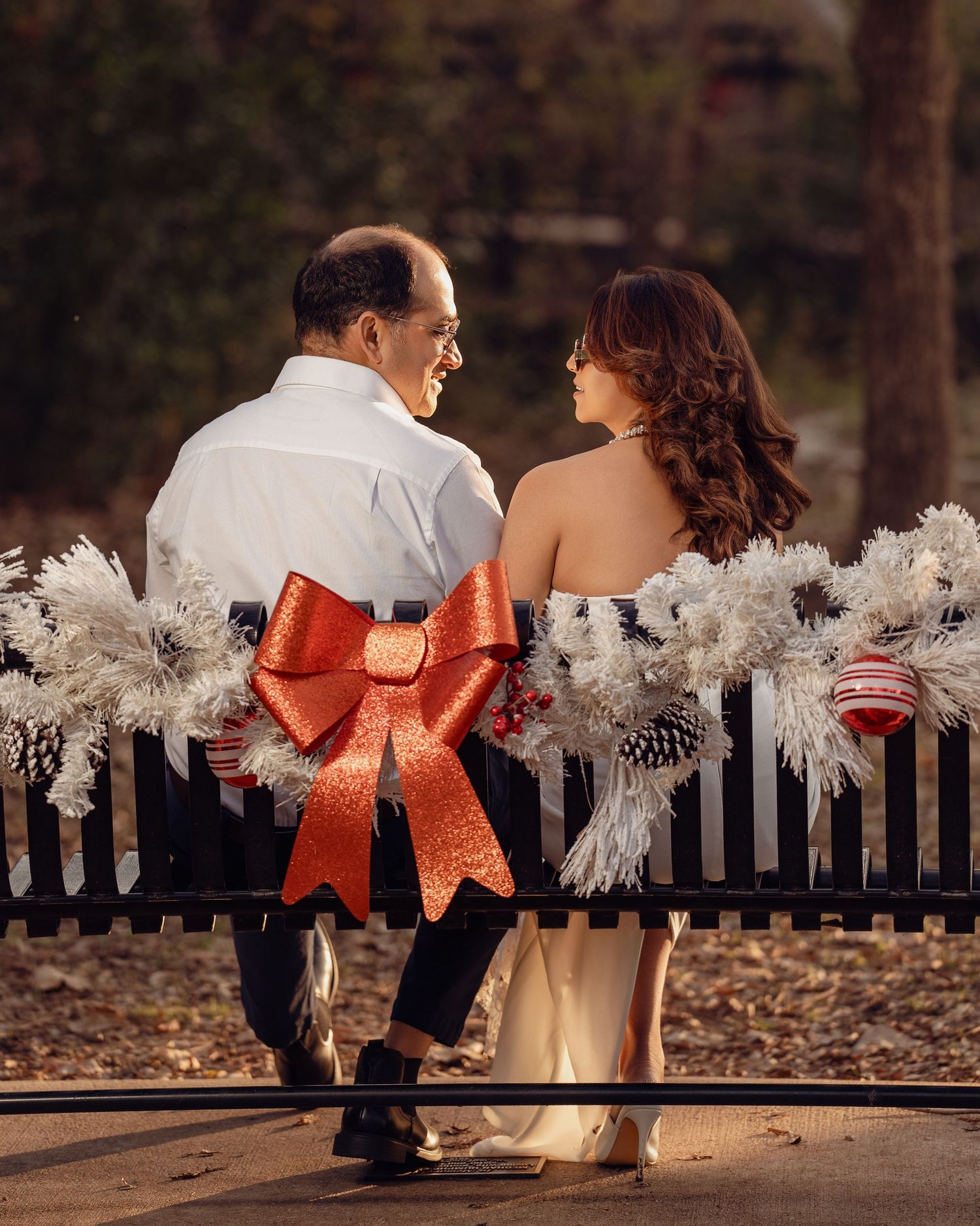 Couple sitting on a Christmas-decorated bench with a red bow, sharing a romantic moment outdoors in warm autumn light.