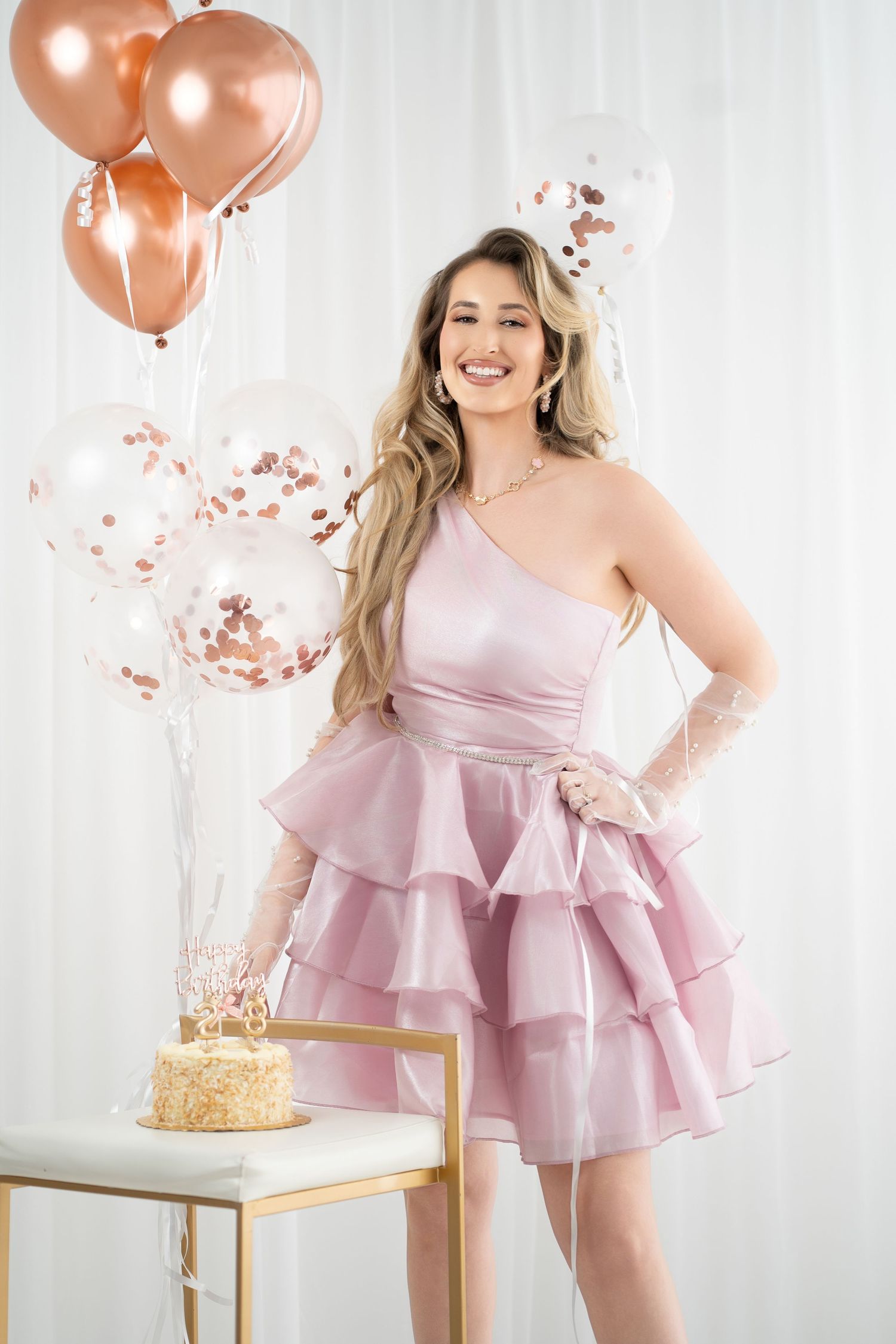 Woman in pink one-shoulder ruffle dress poses with birthday cake and rose gold balloons.