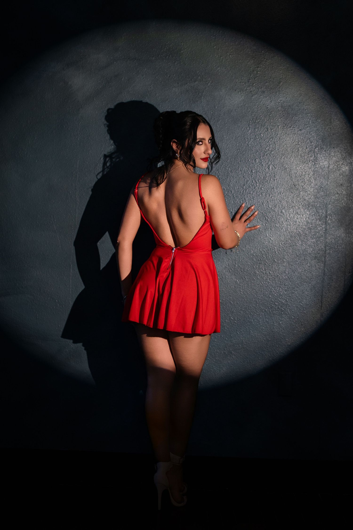 Woman in backless red mini dress poses against dark wall with dramatic spotlight creating striking shadow.