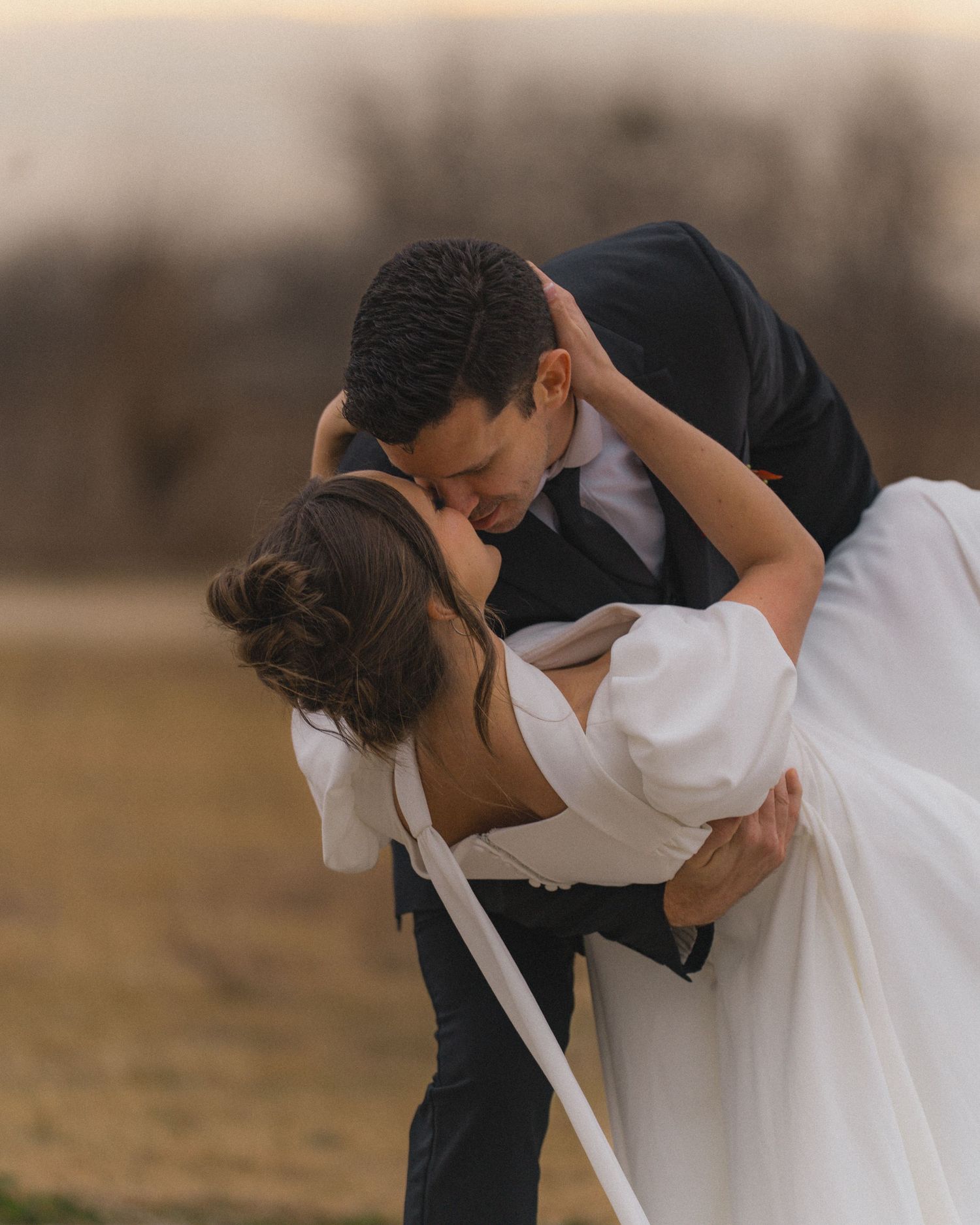 A romantic wedding couple sharing a passionate dip kiss in a rustic outdoor setting at dusk.