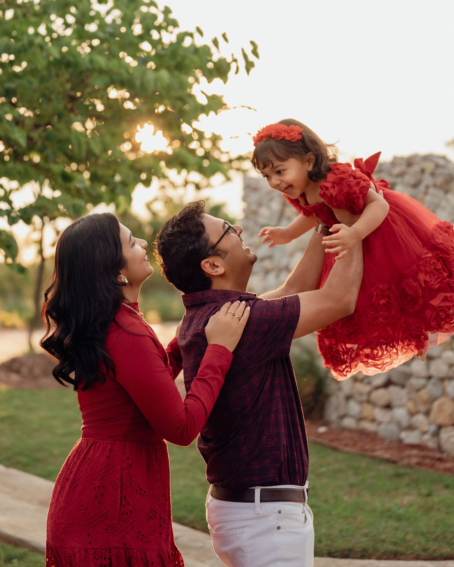 A joyful family in matching red outfits, with a father lifting a smiling toddler girl while mother watches at golden hour.