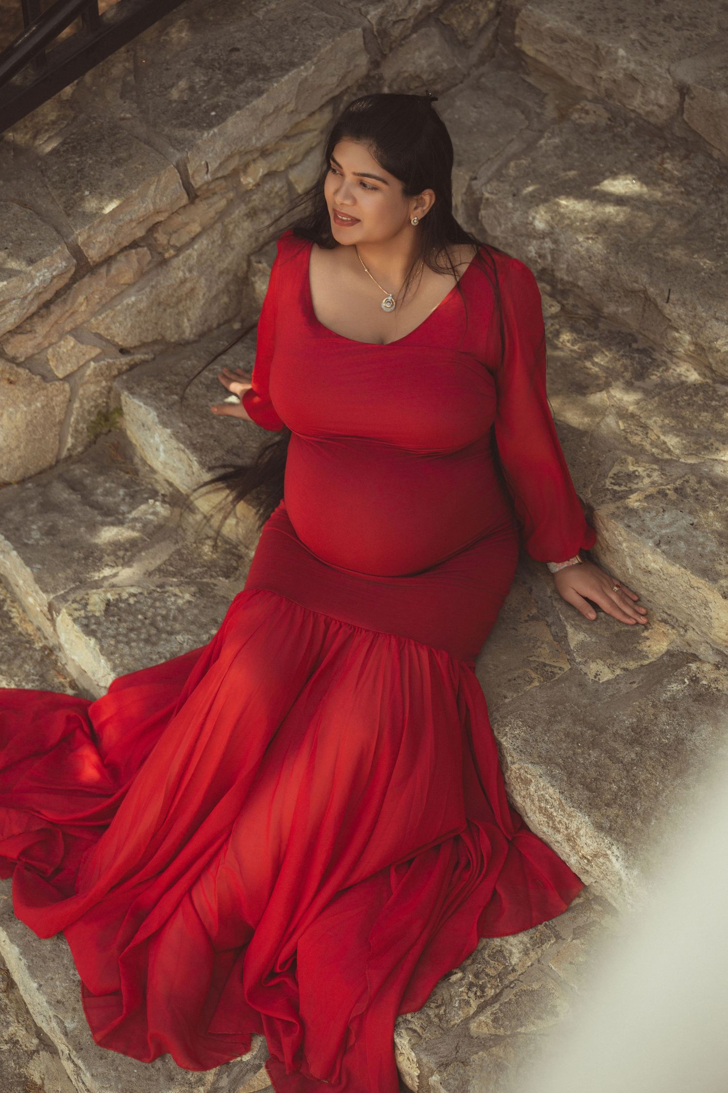 Pregnant woman in elegant red mermaid gown sitting on stone steps, showcasing maternity fashion photography.