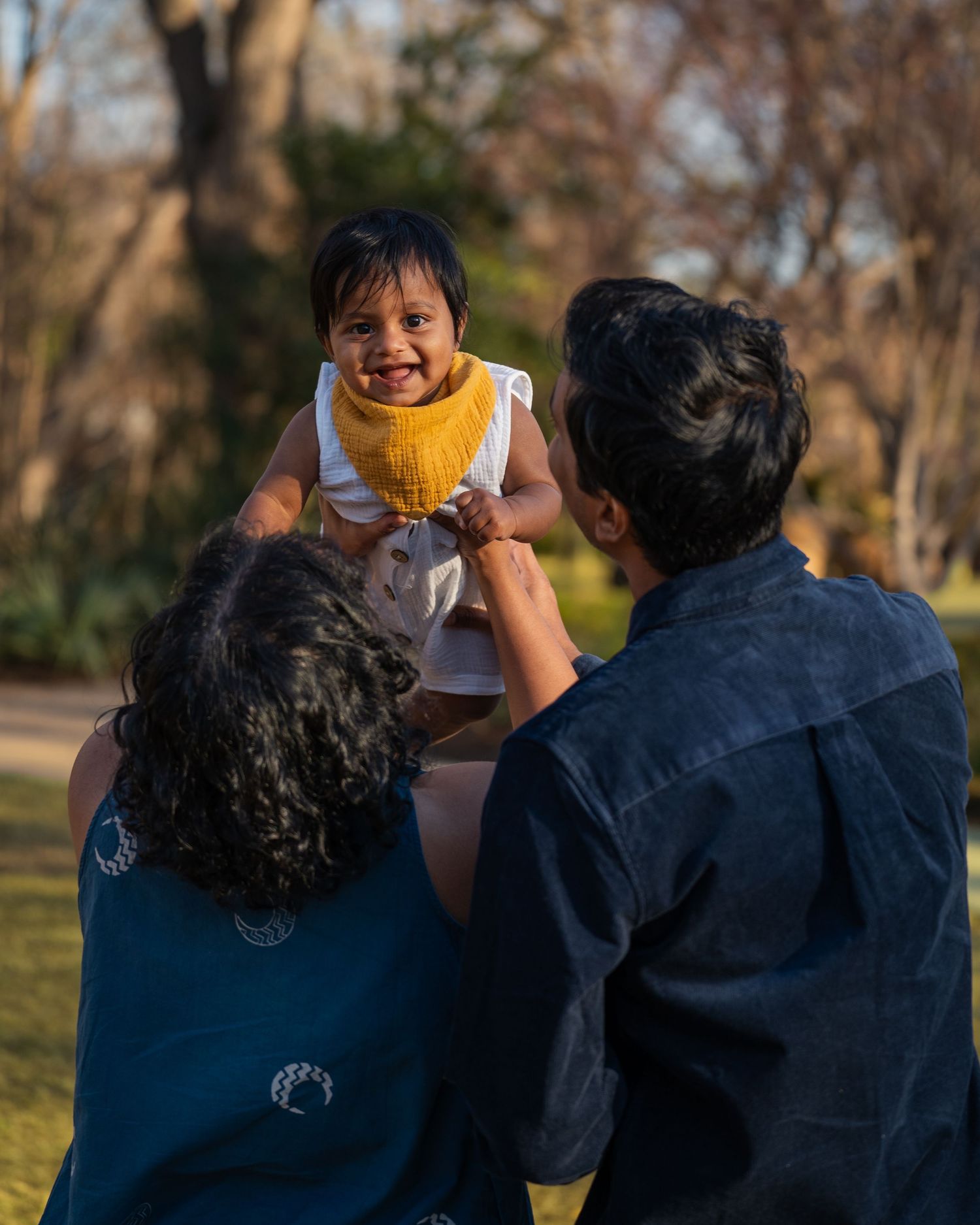 Two parents joyfully lift their smiling baby wearing a yellow bib in a sunny park setting.