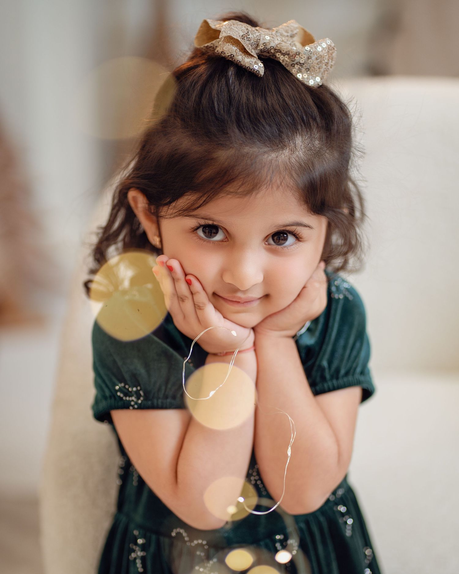 Adorable young girl in green velvet dress with gold bow, posing thoughtfully with bokeh lights in foreground.