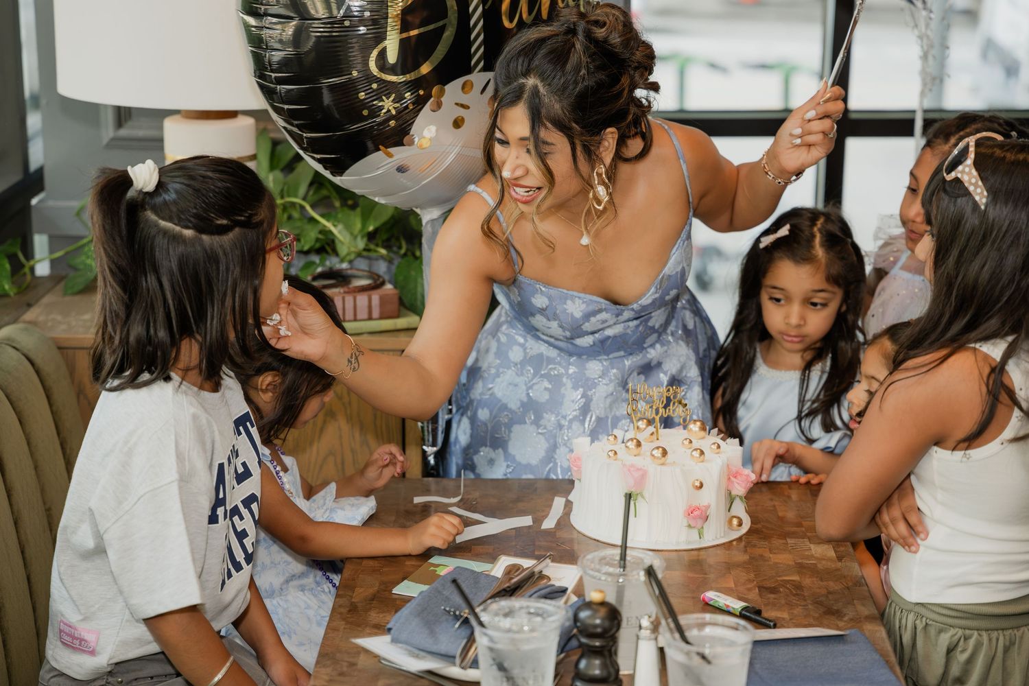 Woman in blue dress celebrates birthday with children around a decorated cake at a restaurant party.