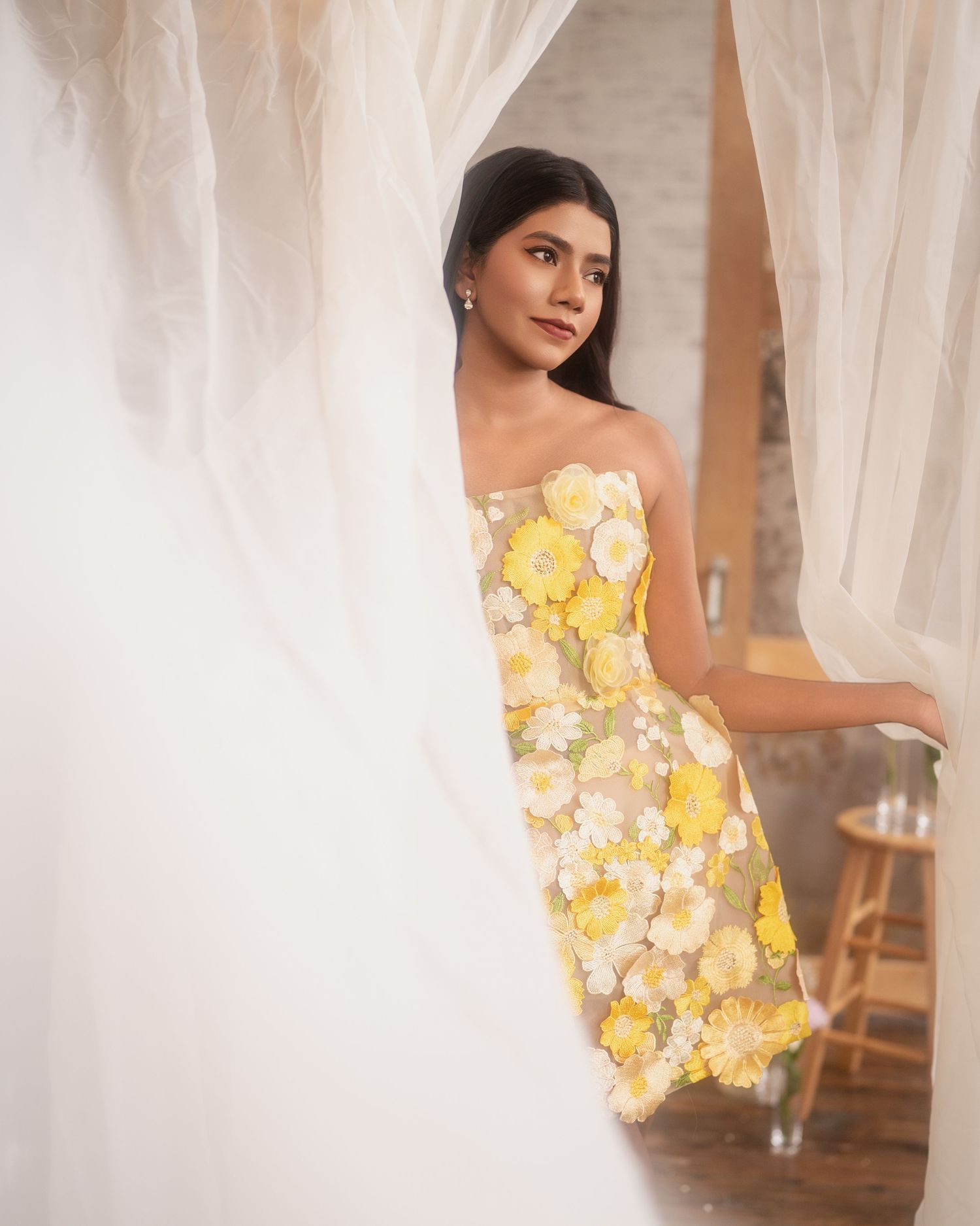 Young woman in a strapless yellow floral dress poses gracefully between sheer white curtains in a bright studio.