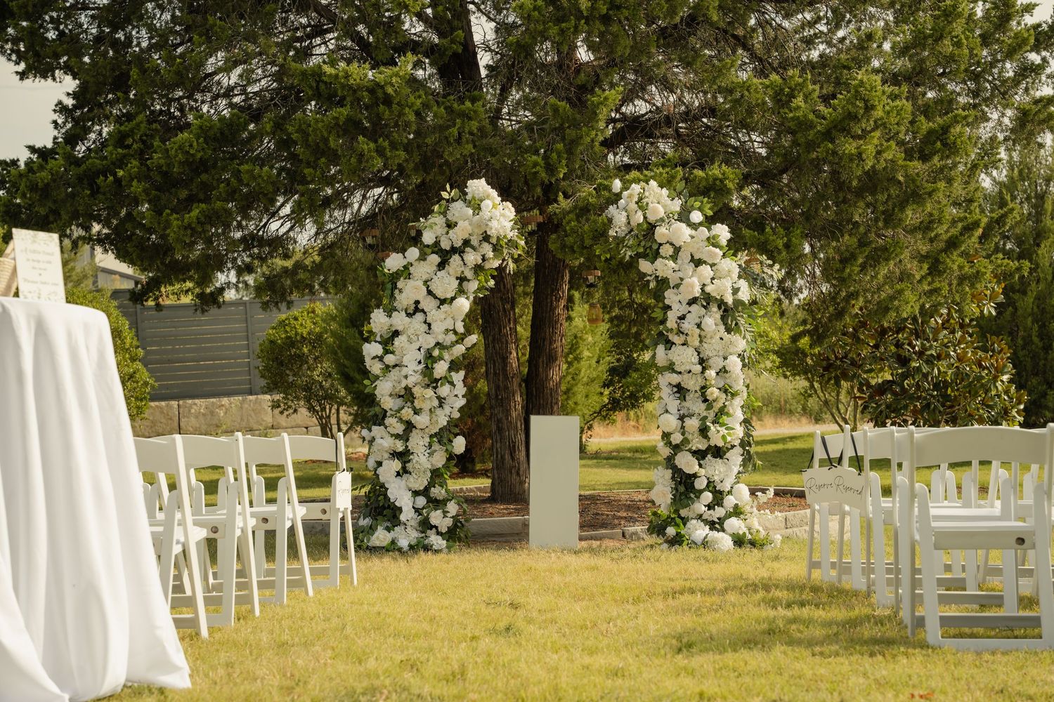 Outdoor wedding ceremony setup with white floral arches, white chairs, and lush green trees in background.