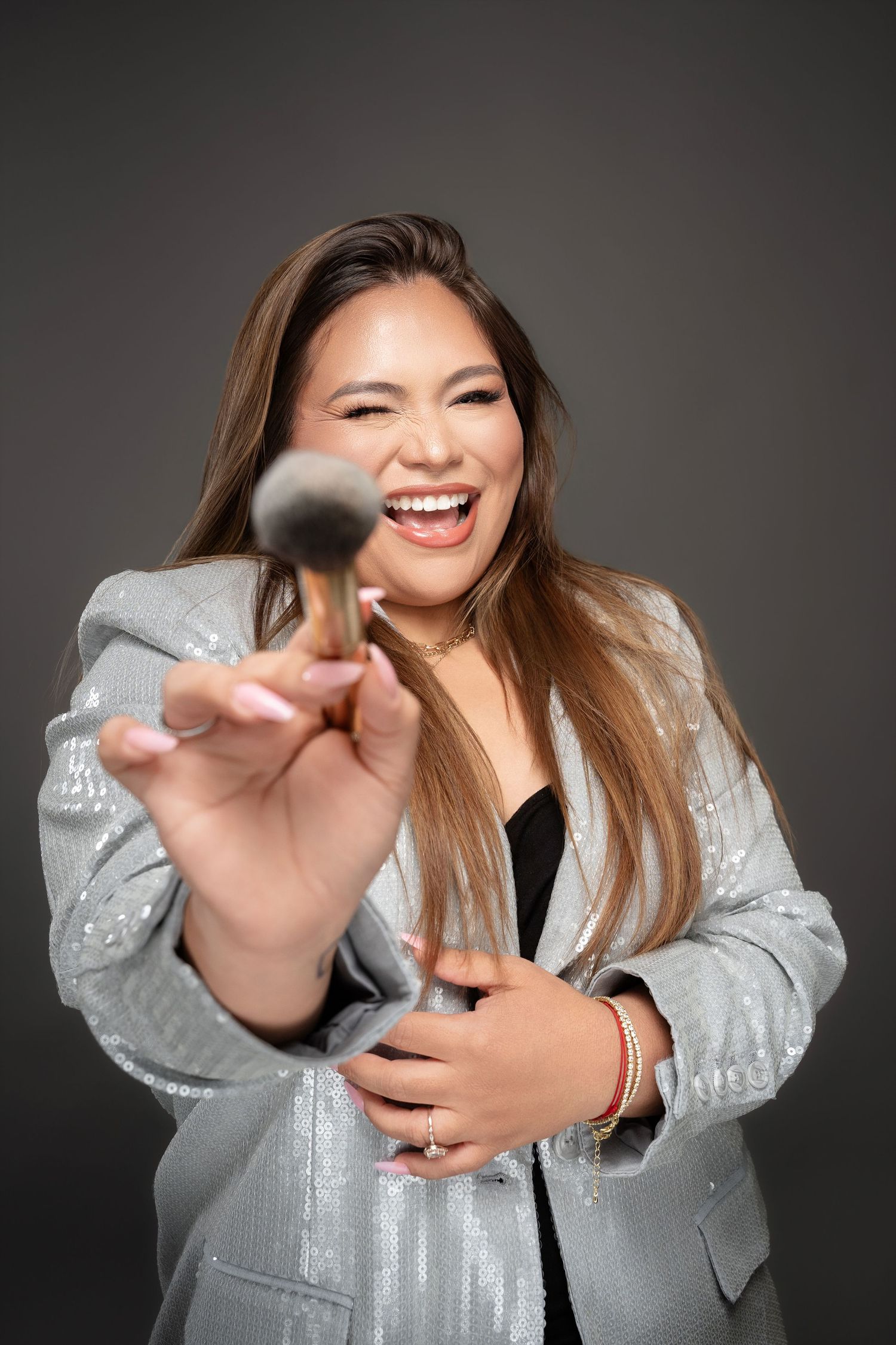 Woman in silver blazer laughing while holding a makeup brush toward camera against gray background.