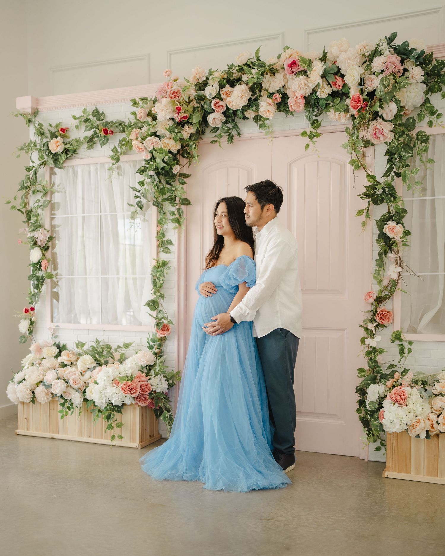 Couple poses under floral arch for maternity photo, woman in blue gown, pink roses and greenery backdrop.