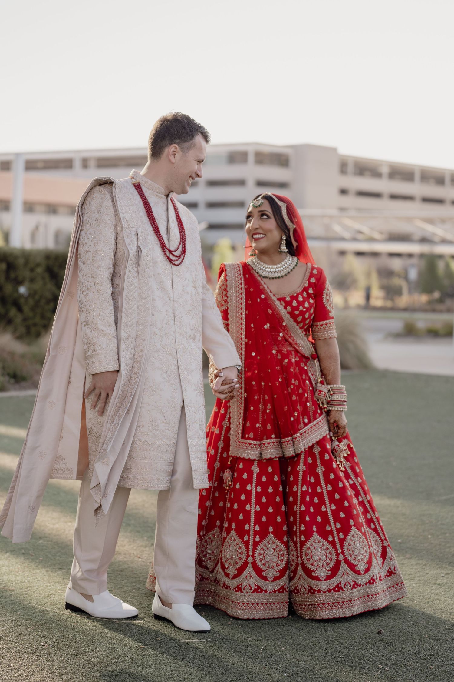 A groom in cream sherwani and bride in red lehenga hold hands smiling at their Indian wedding ceremony outdoors.