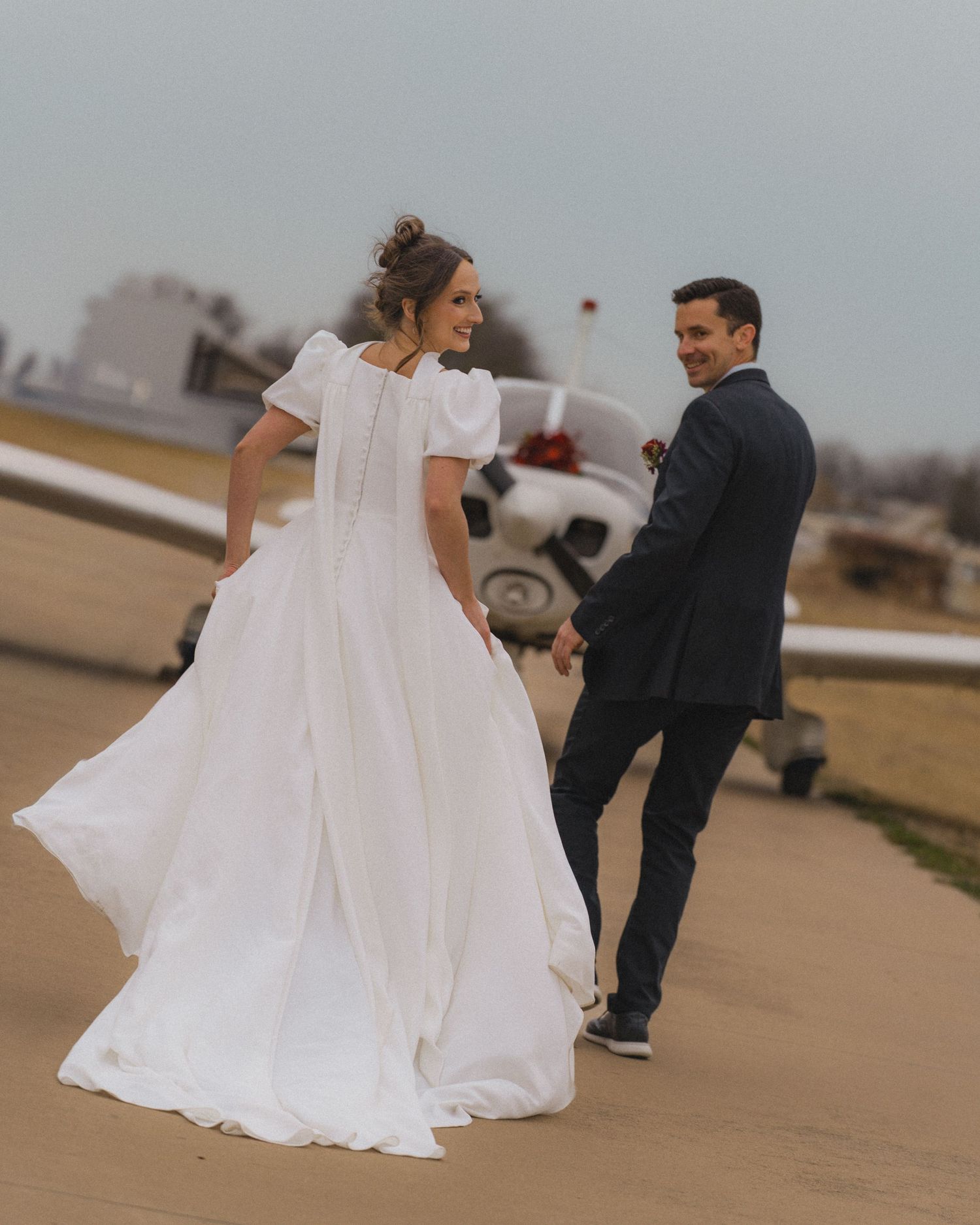 A bride in a flowing white gown and groom in a dark suit walk hand in hand on a runway near a small plane.