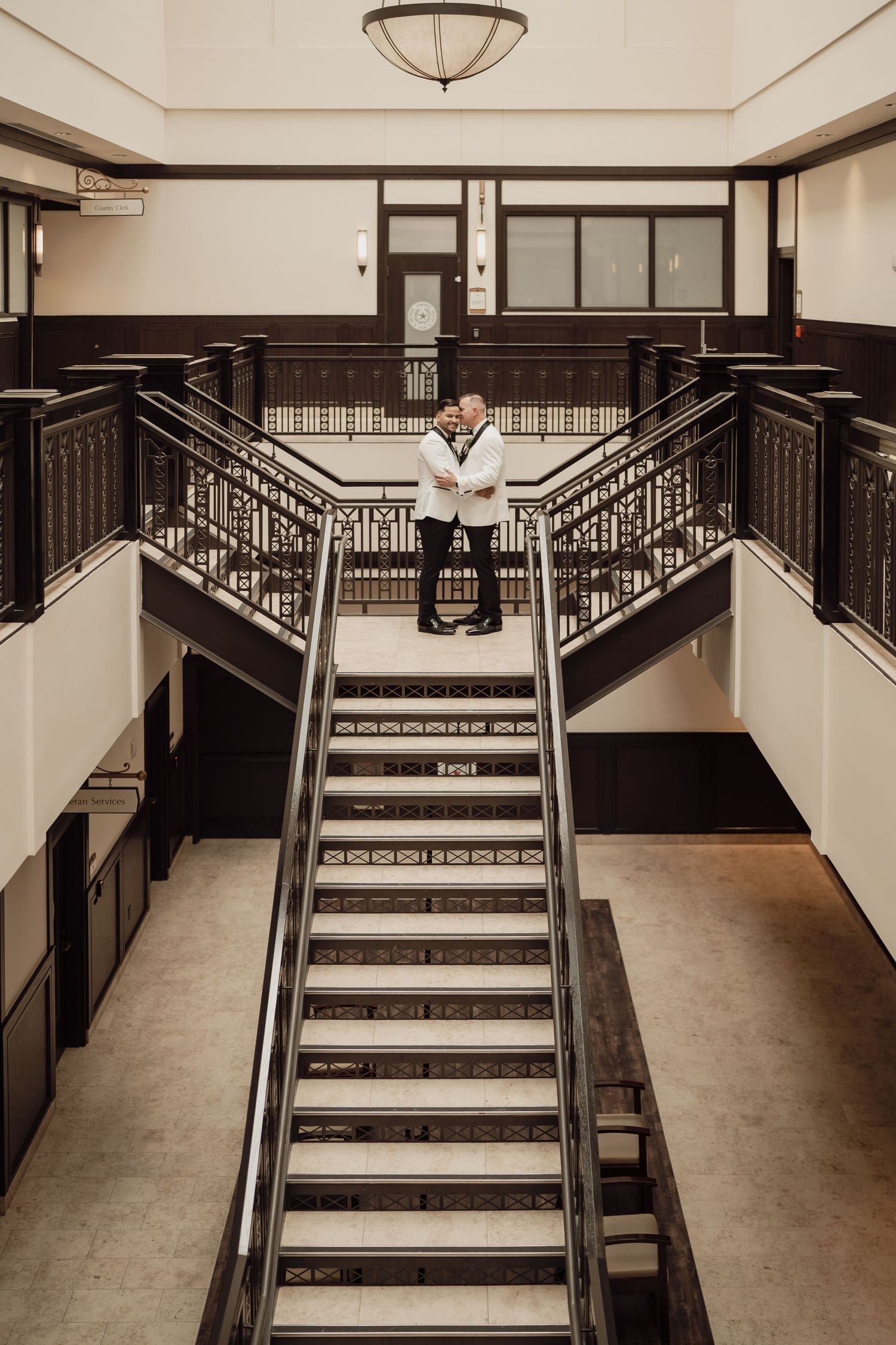 Two people embrace at the top of elegant dark wood staircases in a grand historic building interior.