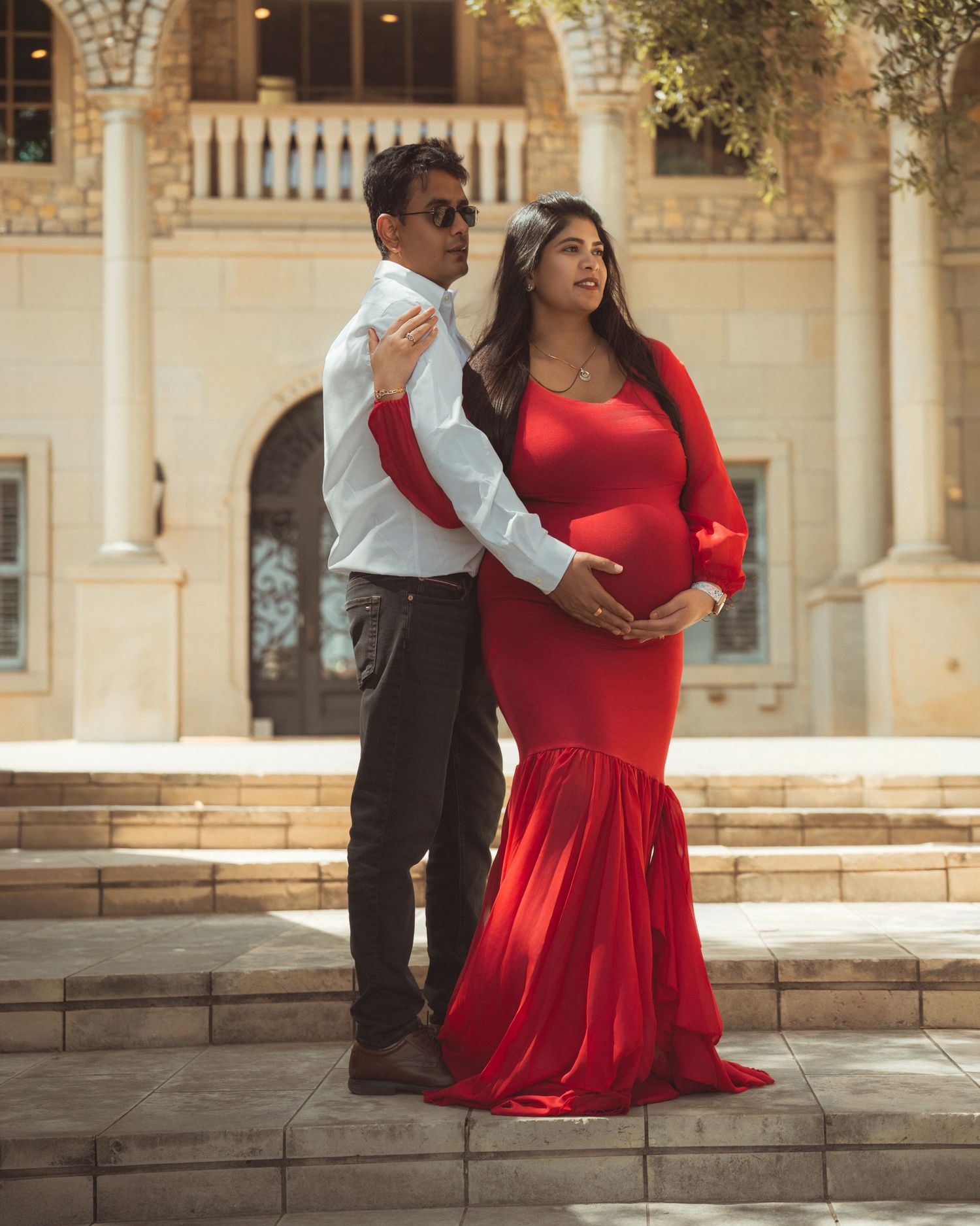 Pregnant woman in red gown poses with partner outside elegant building for maternity photo shoot.
