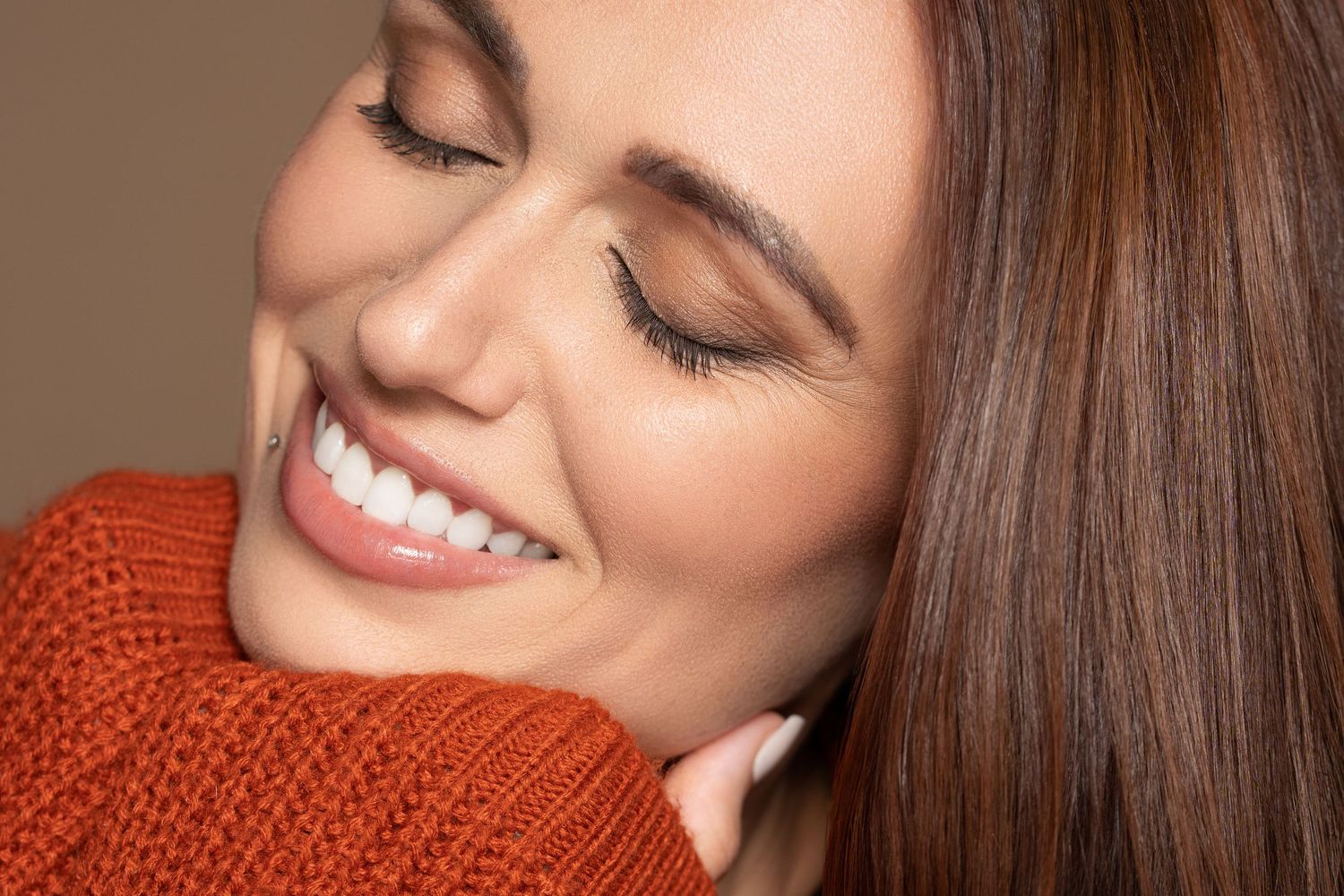 A smiling woman with brown hair wearing an orange sweater, showcasing bright white teeth and glowing skin.