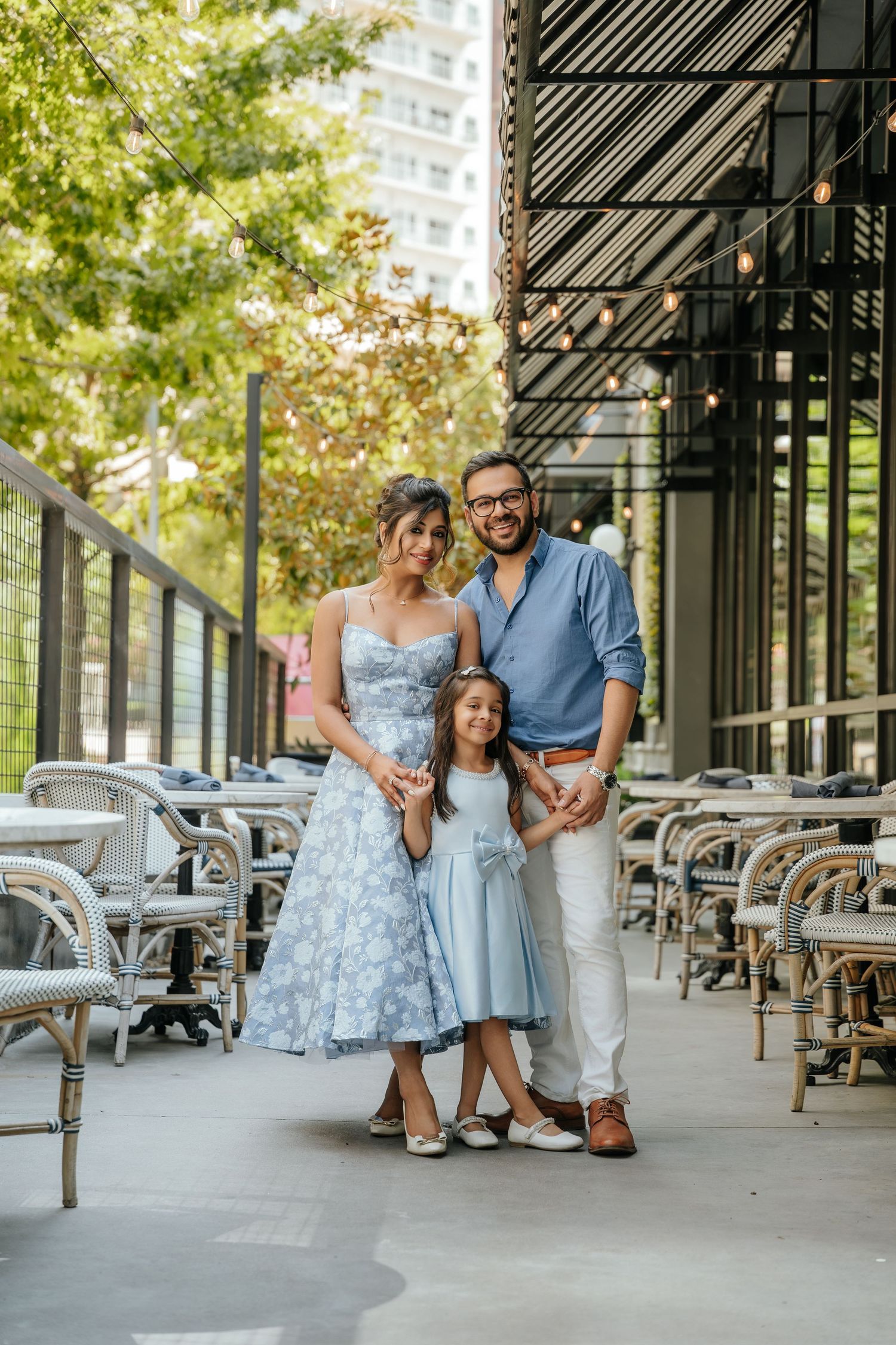 A family of three poses outdoors at a restaurant patio, wearing coordinated blue outfits on a sunny day.