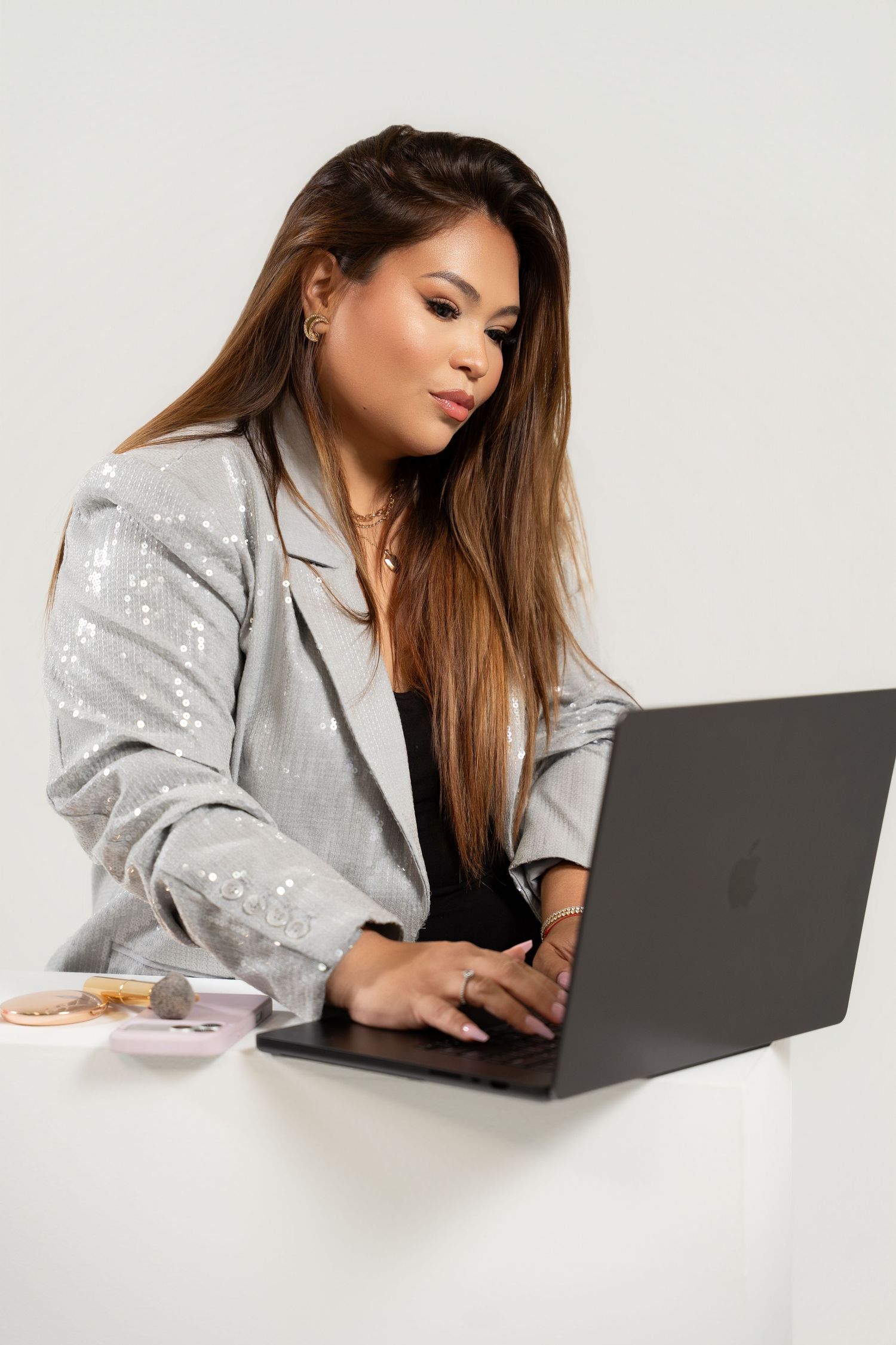 Woman in silver sequin blazer typing on laptop at white desk with makeup items nearby.