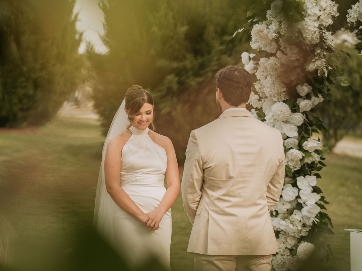 Bride in white halter gown and veil exchanges vows with groom in tan suit under floral arch outdoors.