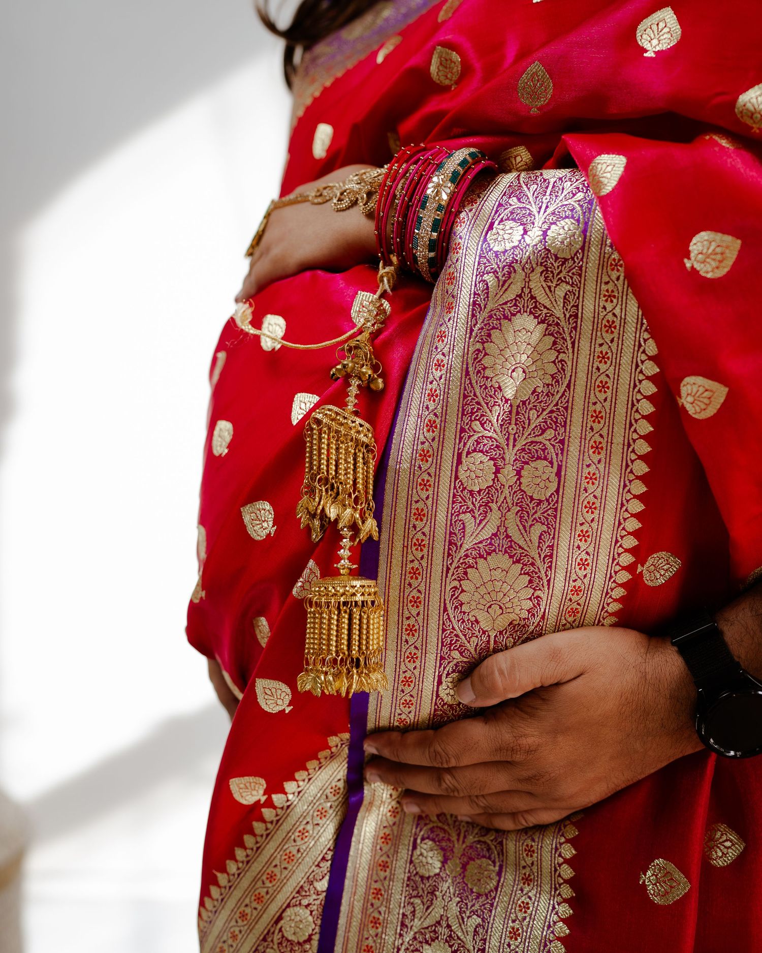 Woman in red and gold Banarasi saree with ornate bangles and traditional gold jhumka charm, elegant Indian bridal attire.