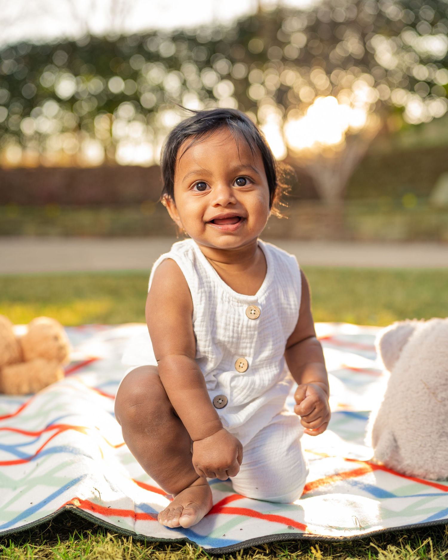 Smiling baby in white romper sitting on colorful blanket outdoors in golden hour sunlight.