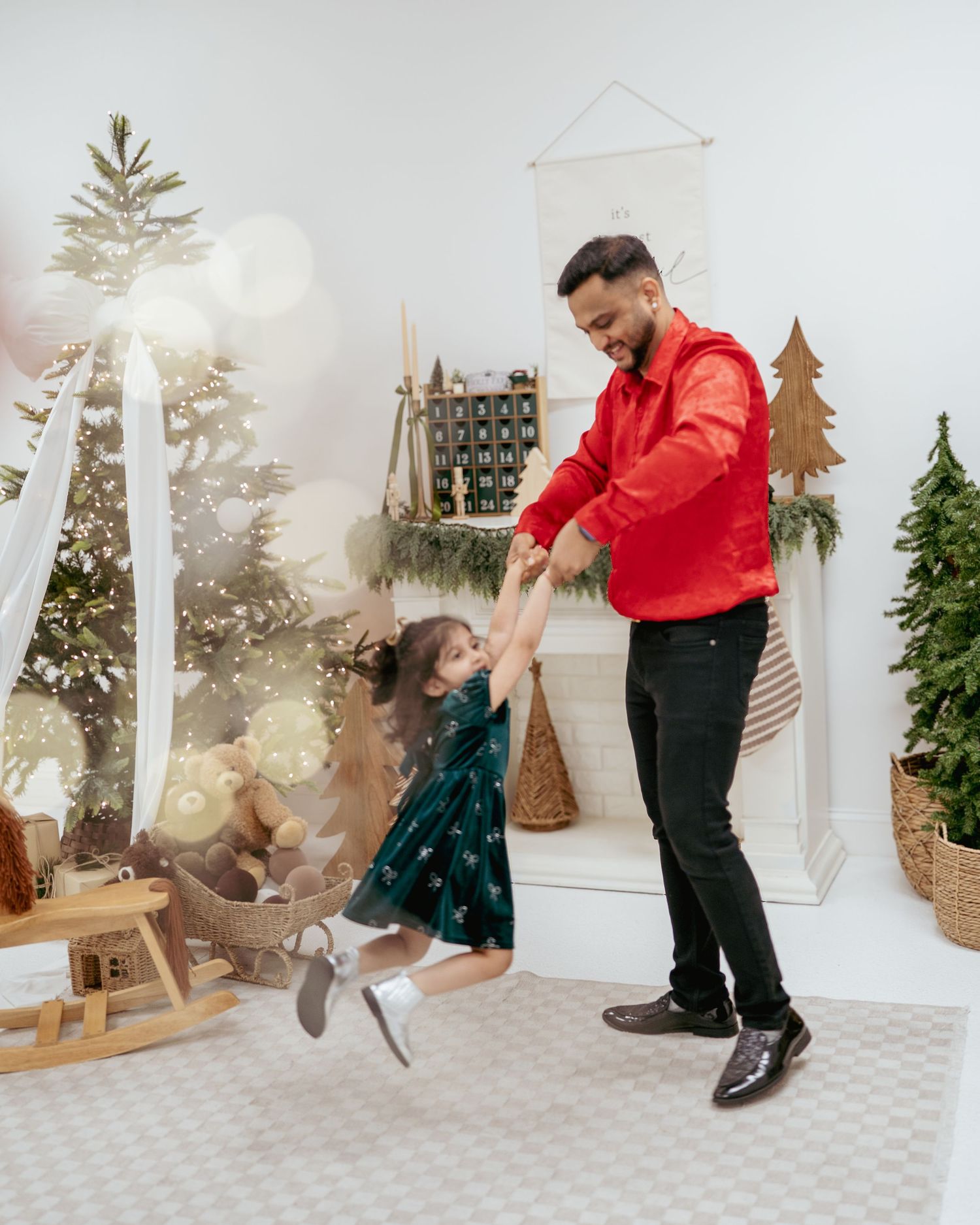 A father in a red shirt joyfully swings his young daughter in a festive Christmas decorated room.