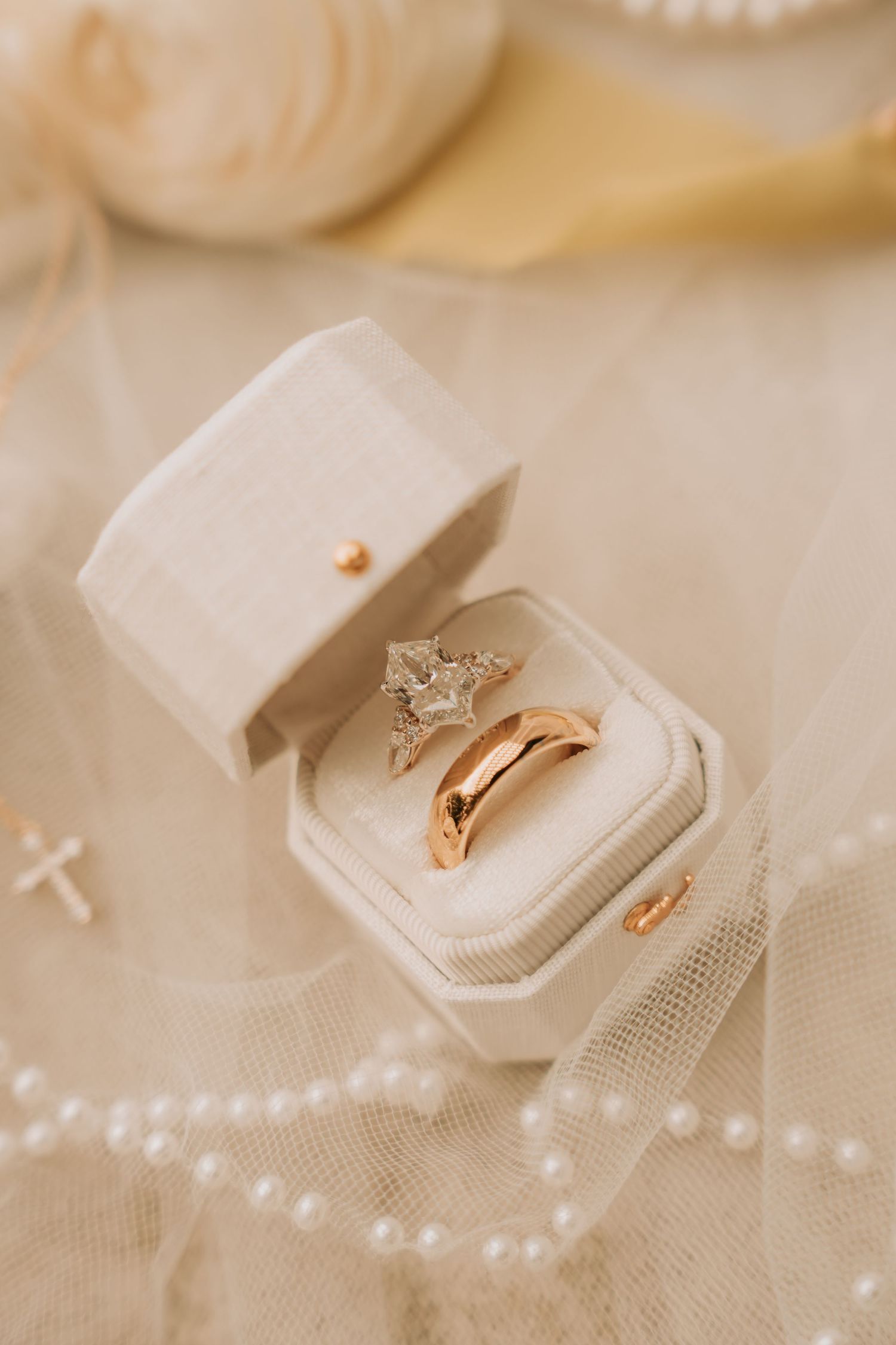 Two gold wedding rings in a cream velvet ring box, with pearl accents and roses on a soft tulle background.