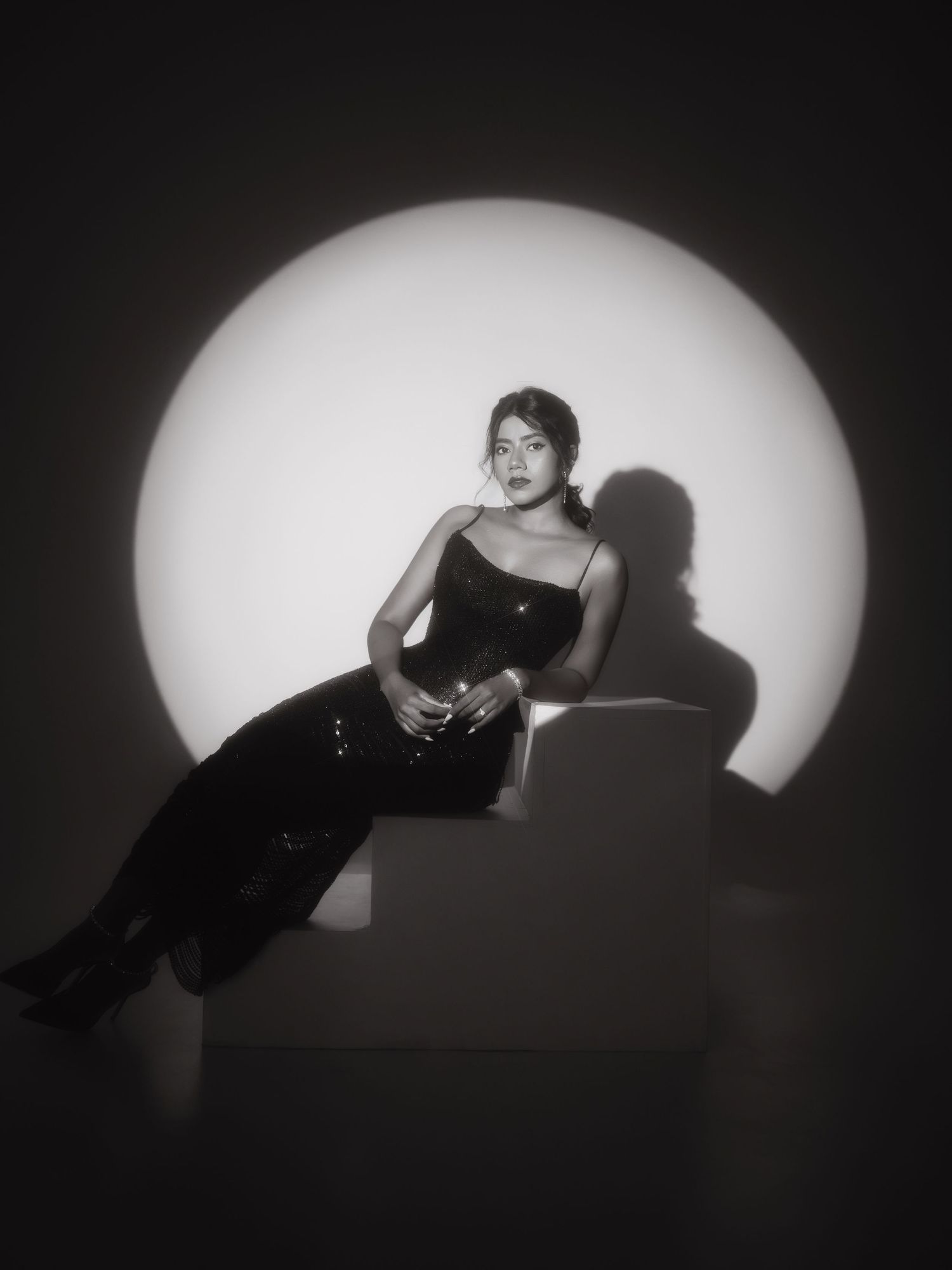 Woman in black sequin dress poses on white cube against circular light backdrop in dramatic black and white photo.