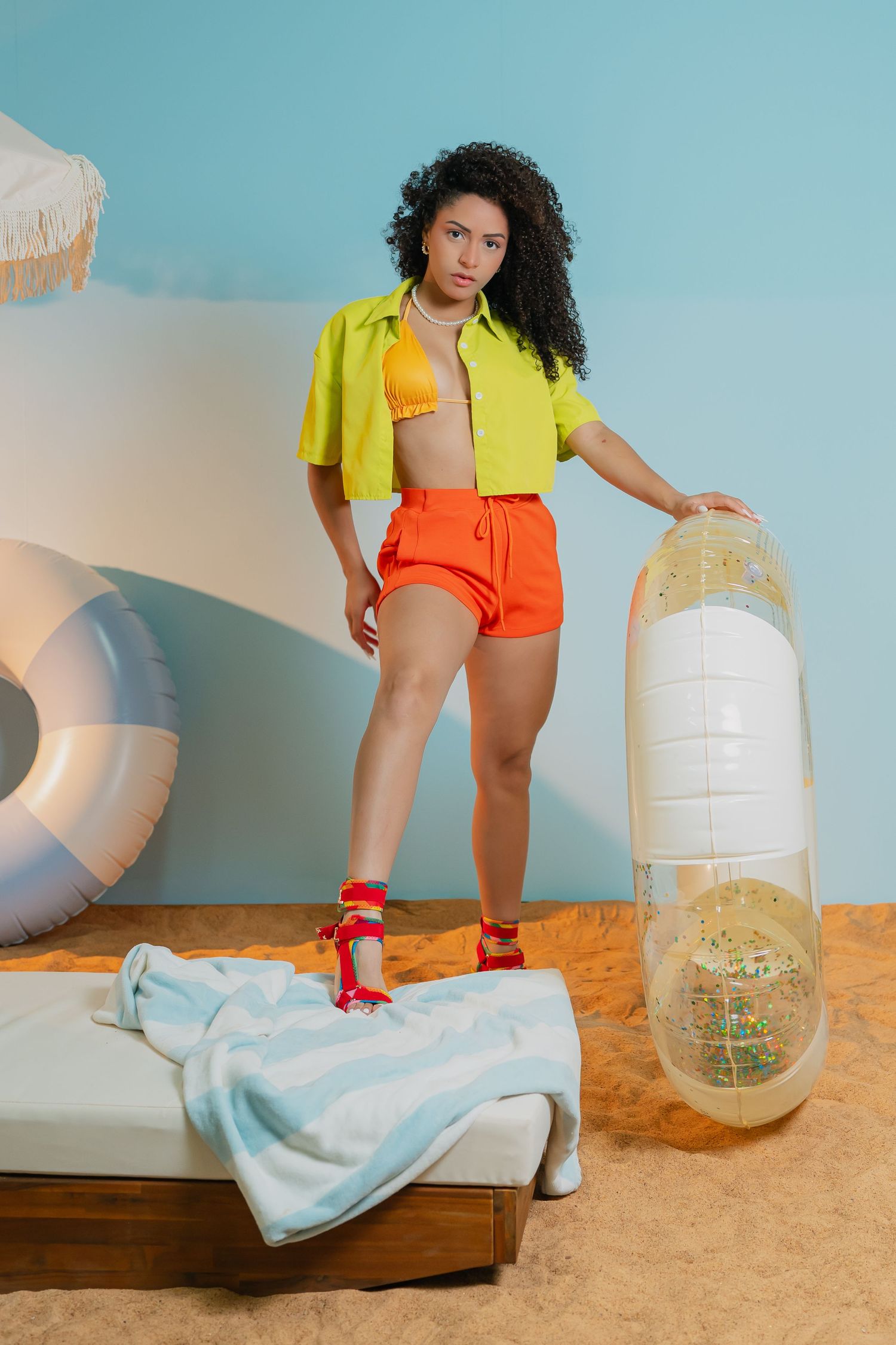Woman in yellow crop top and orange shorts poses with inflatable pool float on sandy beach studio set.