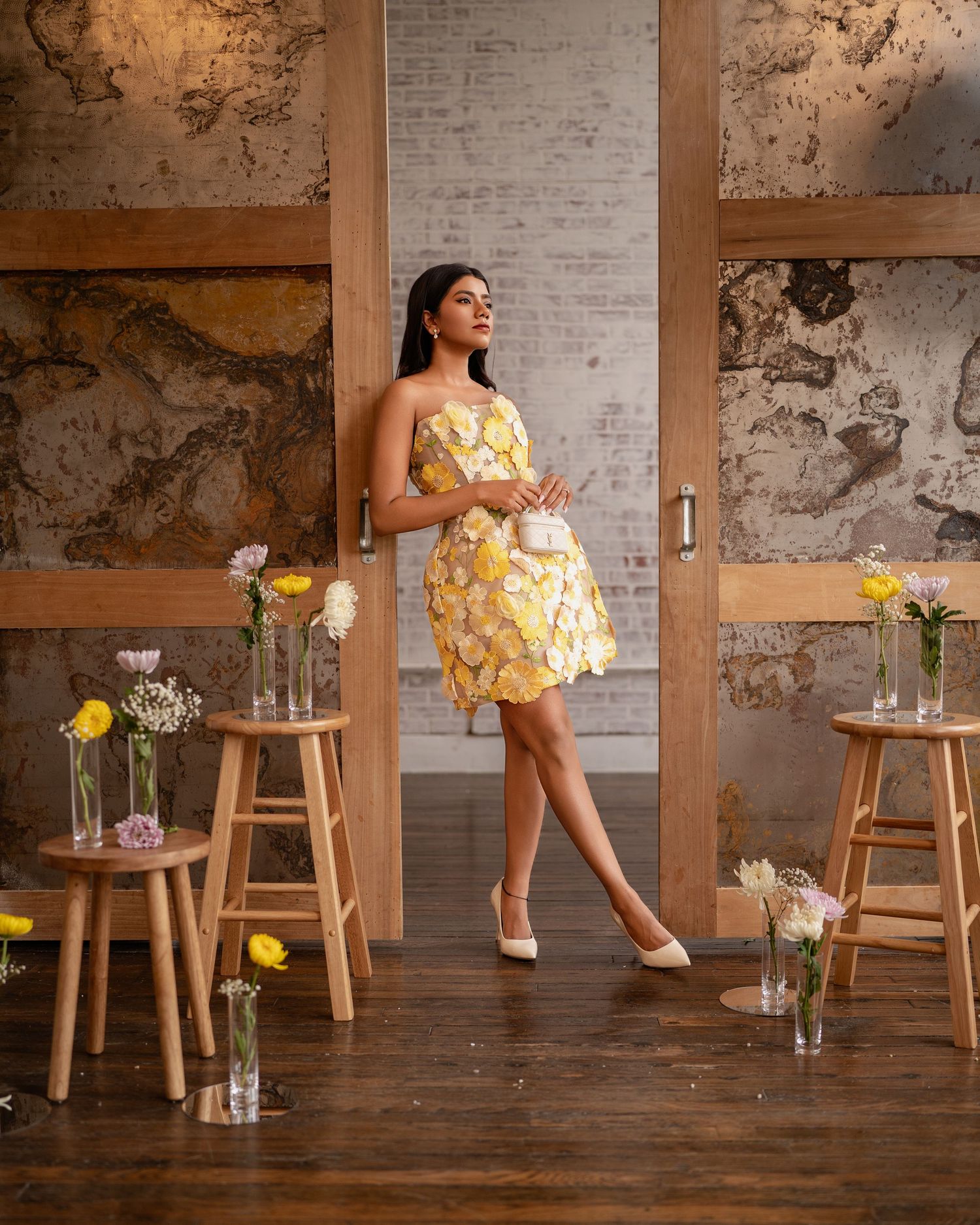Woman in yellow floral mini dress poses among wooden stools with flower vases in a rustic studio setting.