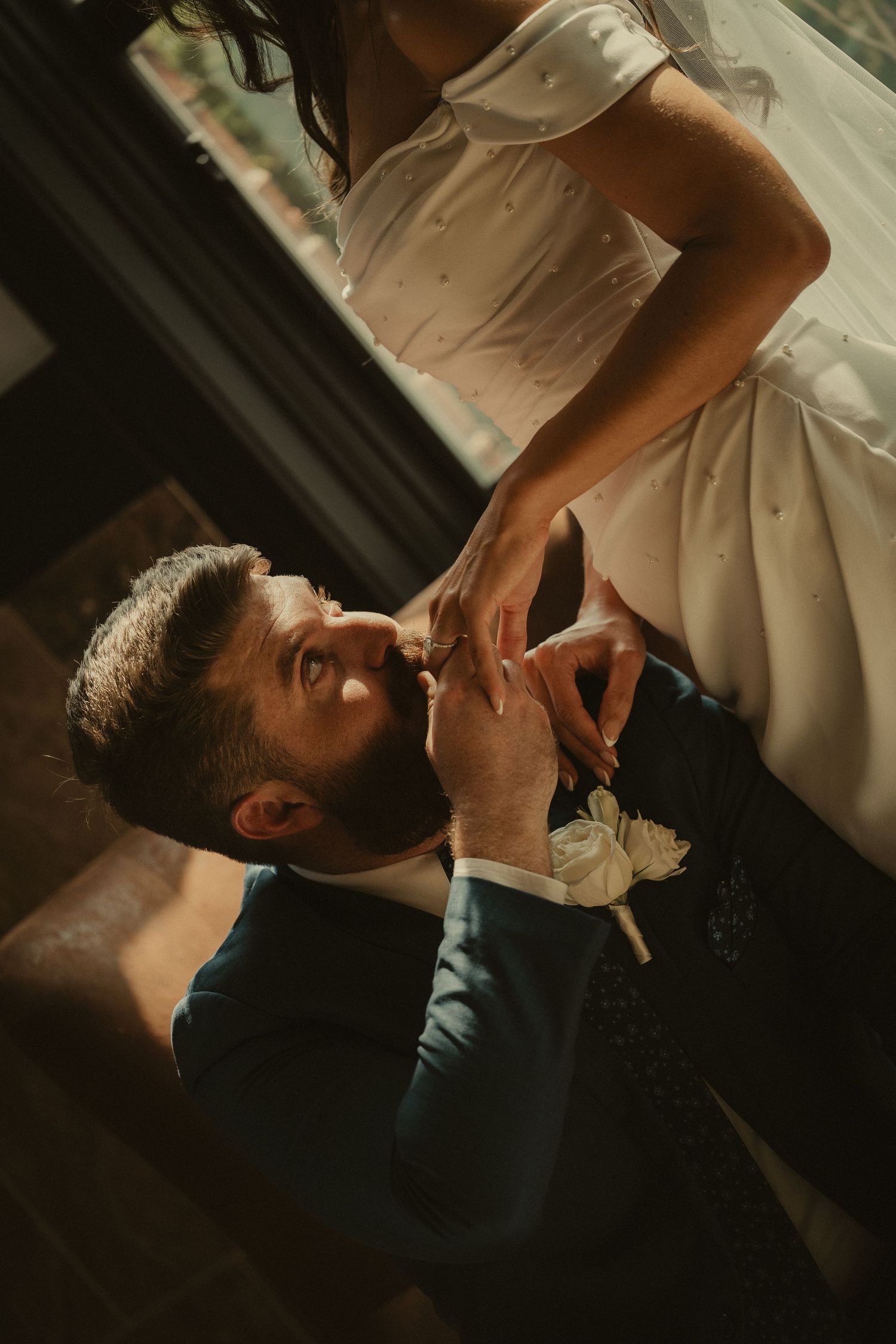 Groom in blue suit kisses bride's hand tenderly during romantic wedding moment indoors with warm lighting.