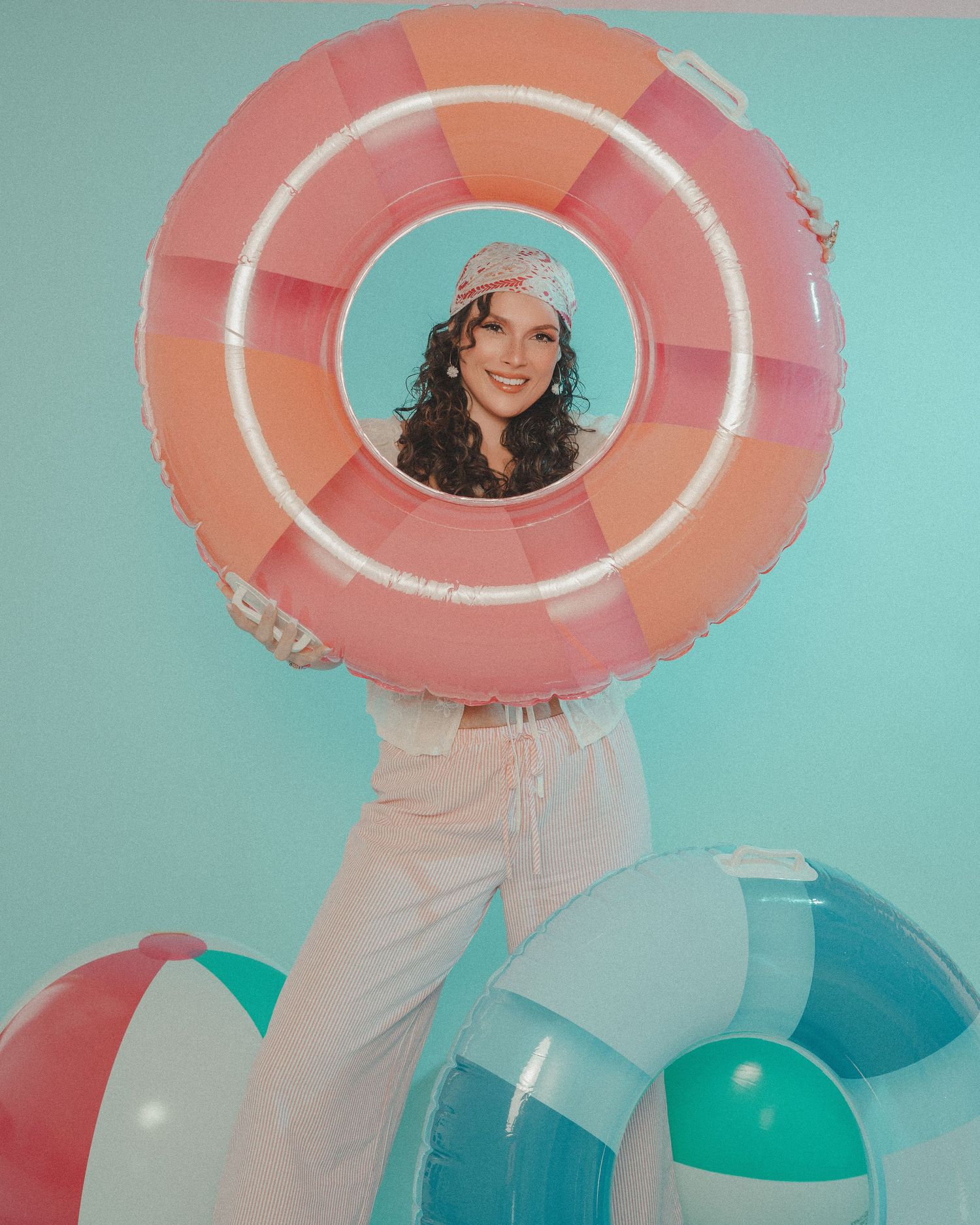 Smiling woman posing behind a large pink inflatable pool ring with beach balls on teal background.