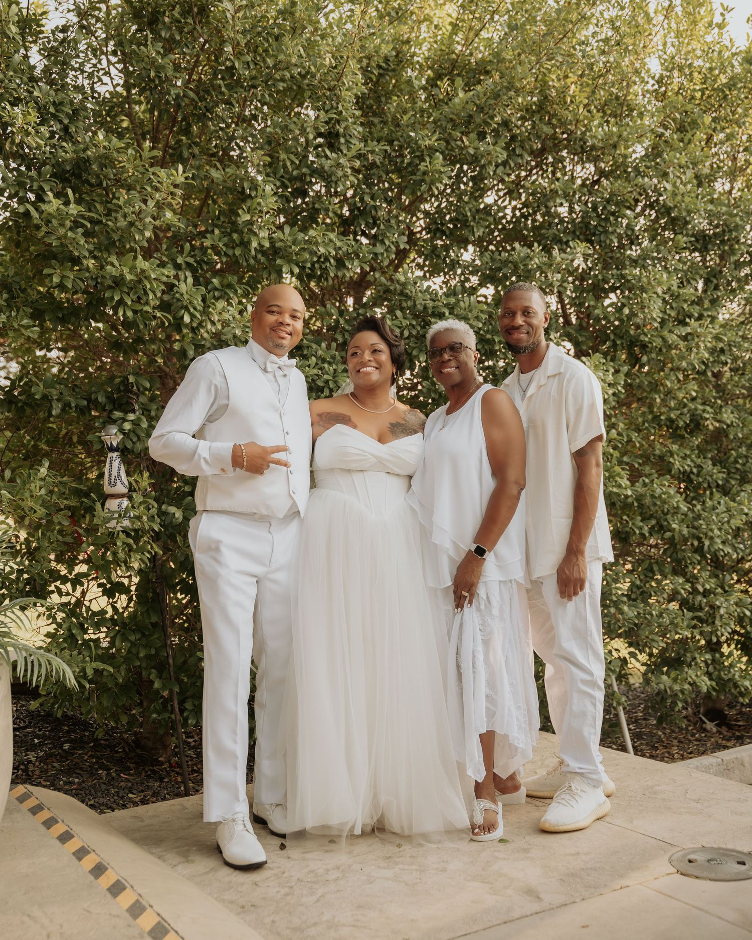 Four people in white outfits posing together outdoors, including a bride in a tulle gown at a wedding celebration.