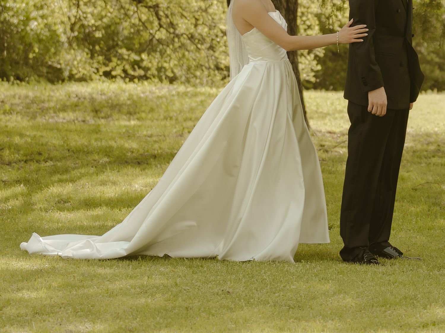 Bride in white wedding gown with train stands beside groom in black suit on green grass outdoors.