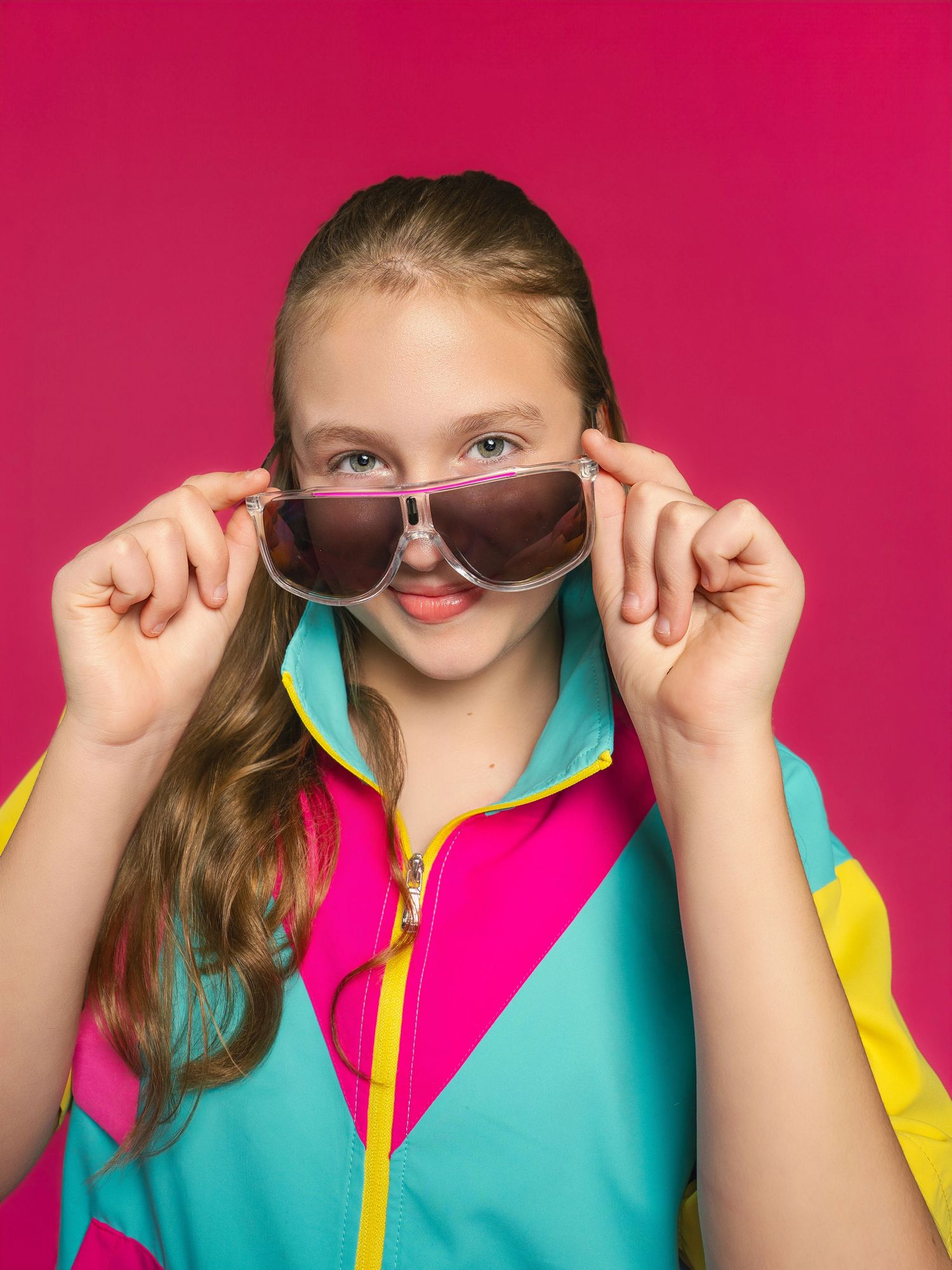 A young girl in a colorful 80s-style tracksuit holds oversized sunglasses against a bright pink background.