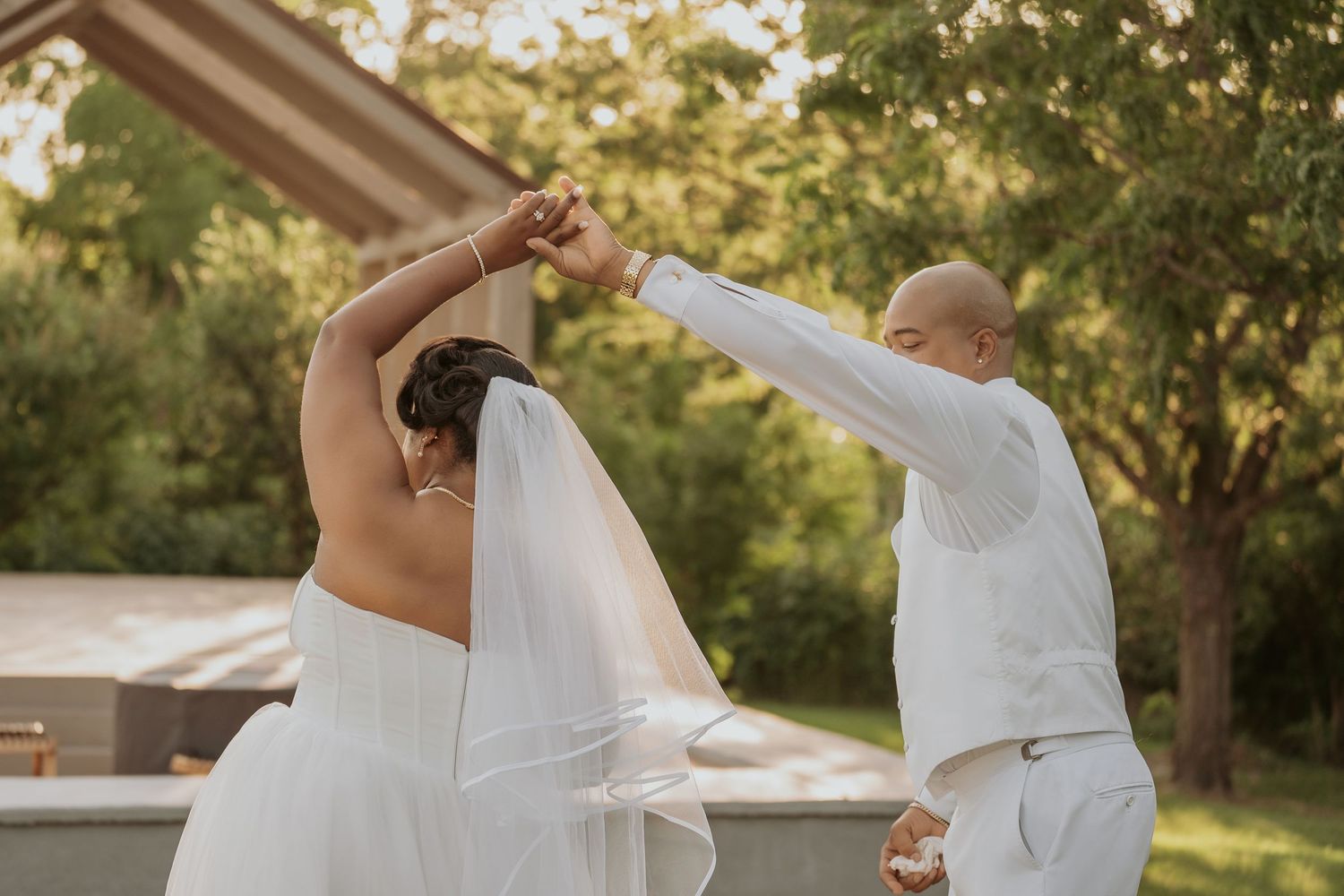 A bride and groom share a romantic dance outdoors, twirling in white wedding attire surrounded by lush greenery.