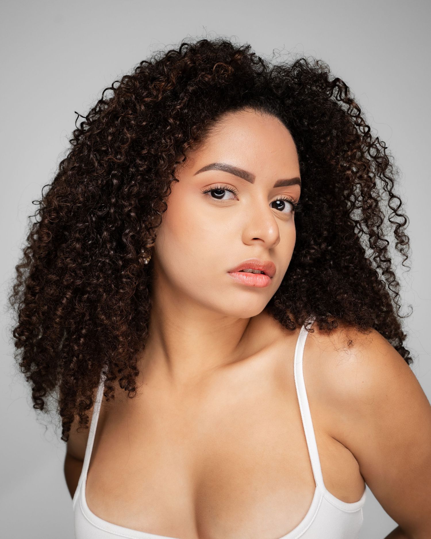 Young woman with voluminous curly dark hair wearing a white top, posing against a light gray background.