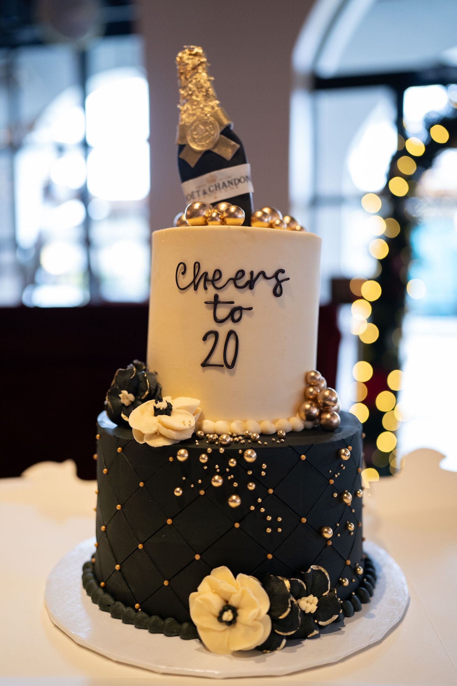 Elegant two-tier black and white celebration cake decorated with gold accents and floral details against a bokeh background.