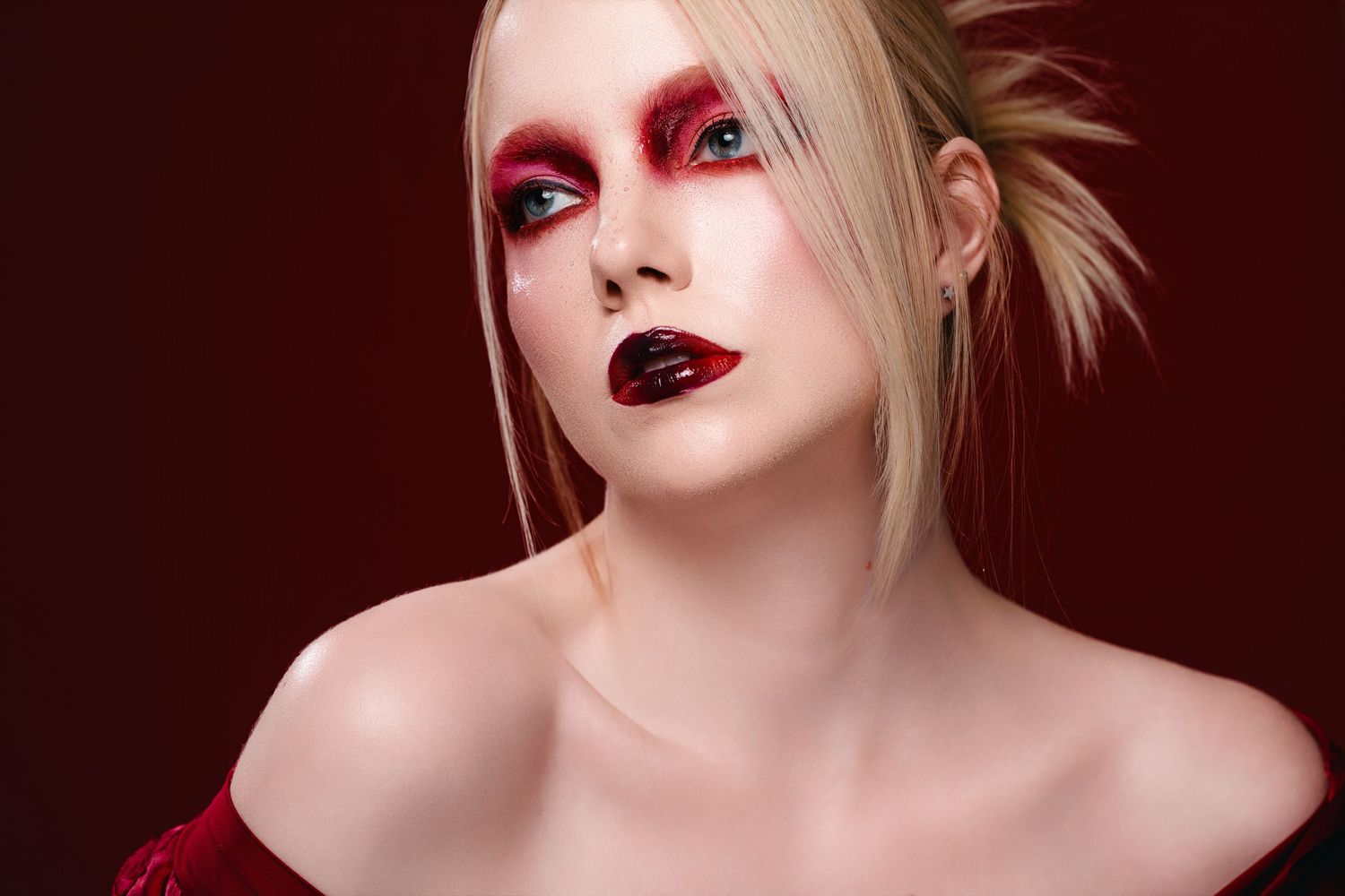 Woman with bold red and black makeup, blonde hair, and bare shoulders poses against a dark red background.
