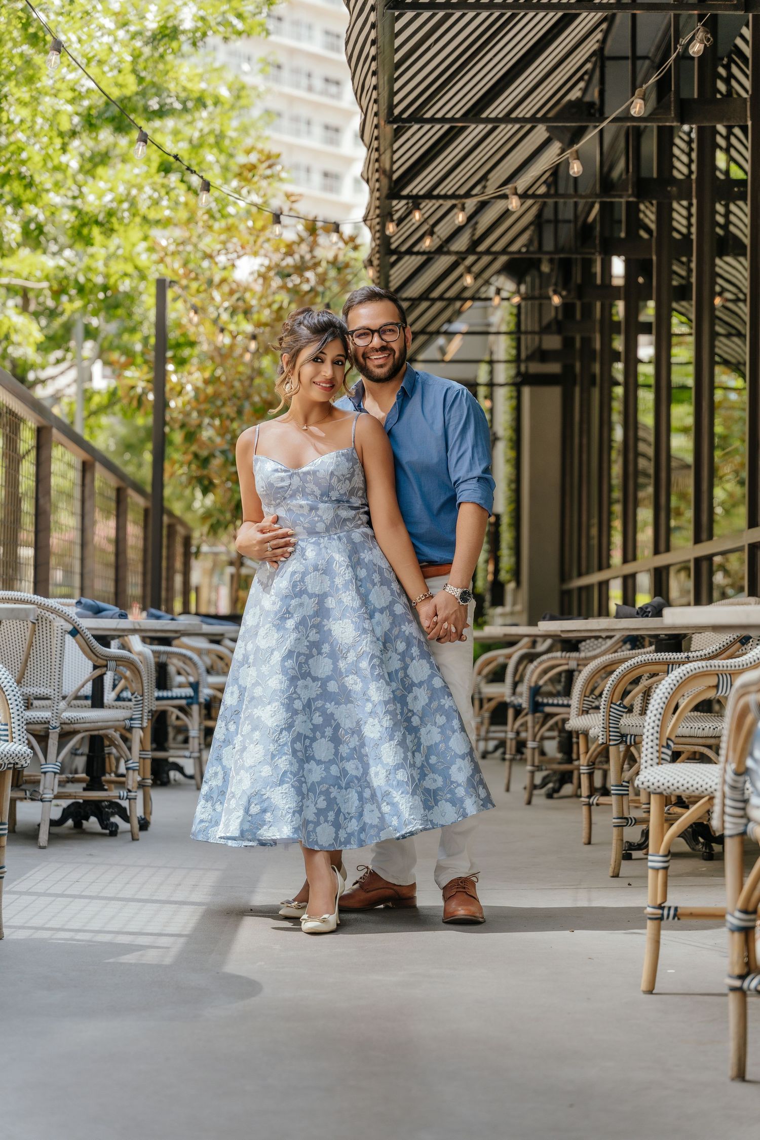 Smiling couple posing on a restaurant patio, woman in blue floral dress, man in blue shirt, holding hands.