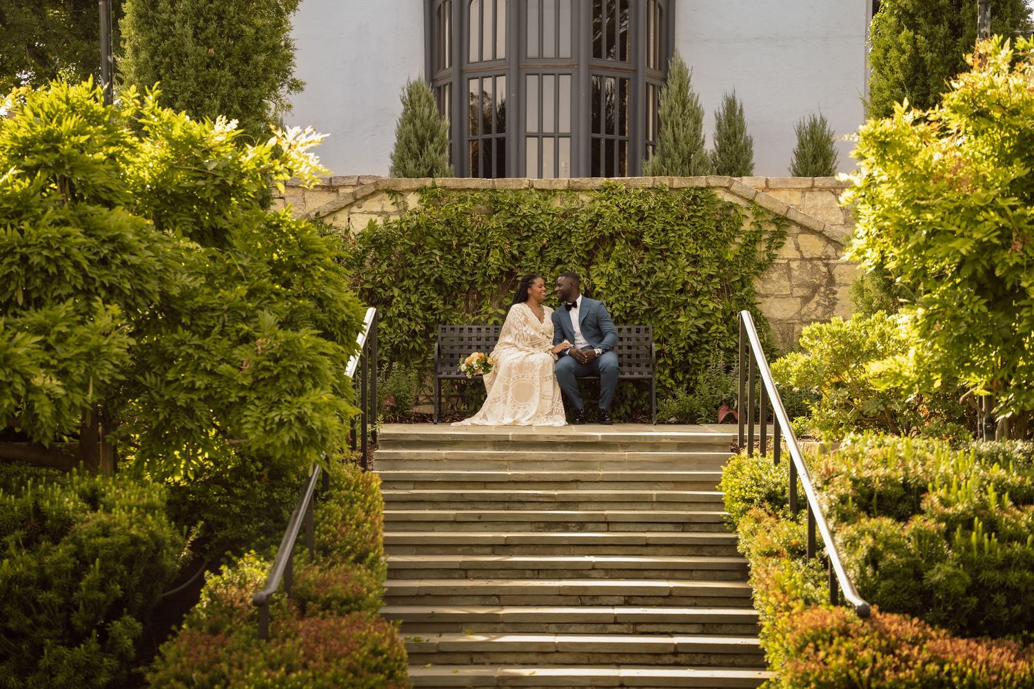 Bride and groom pose on stone steps surrounded by lush greenery outside an elegant building on their wedding day.