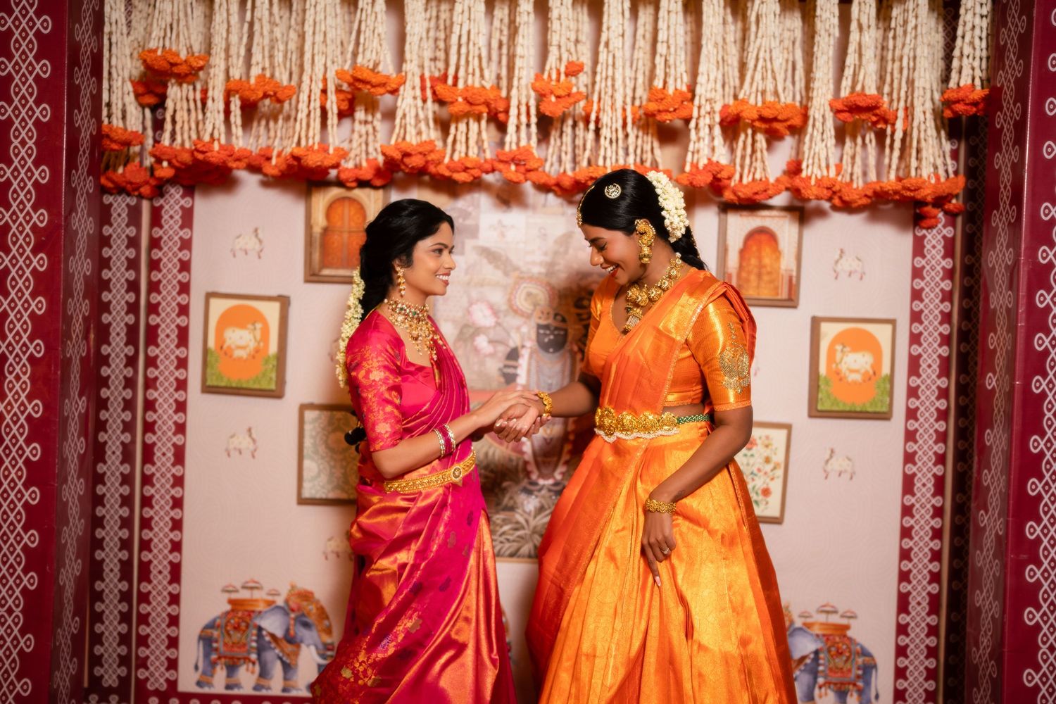 Two women in vibrant silk sarees exchange gifts at a traditional South Indian ceremony with floral decorations.