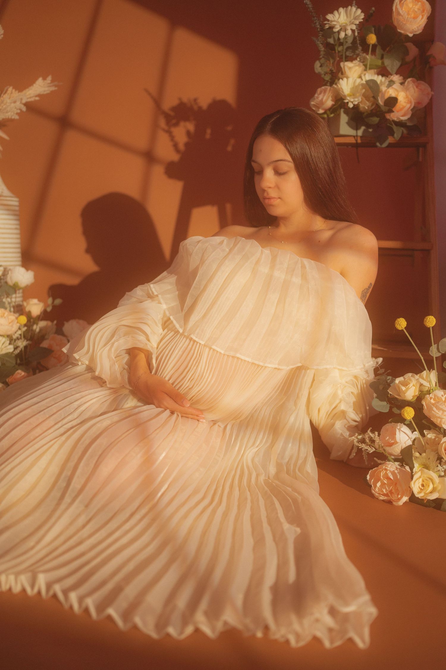 Pregnant woman in white off-shoulder pleated dress sitting among flowers in warm terracotta-toned studio.