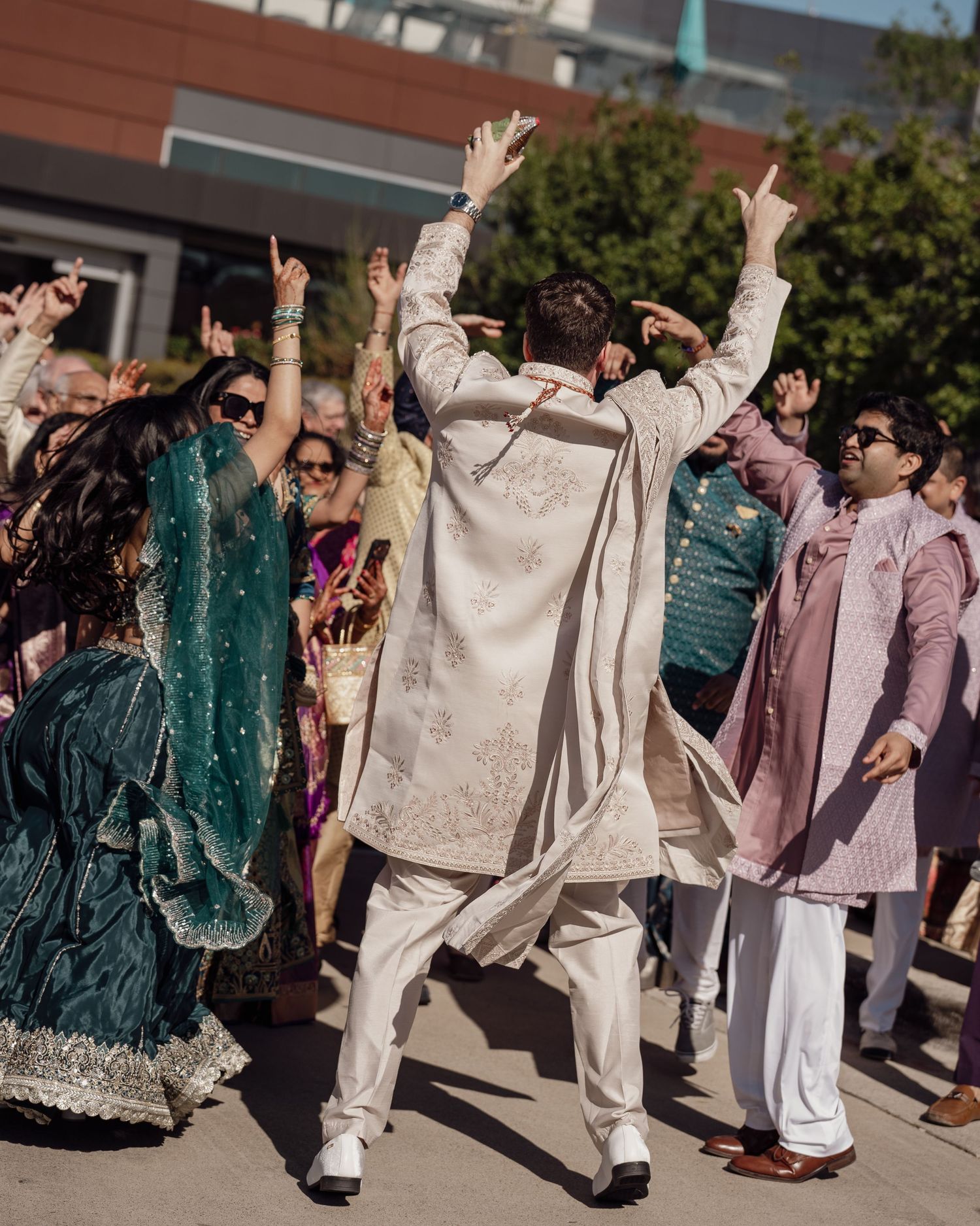 Guests in vibrant Indian attire dance joyfully outdoors at a traditional wedding celebration.