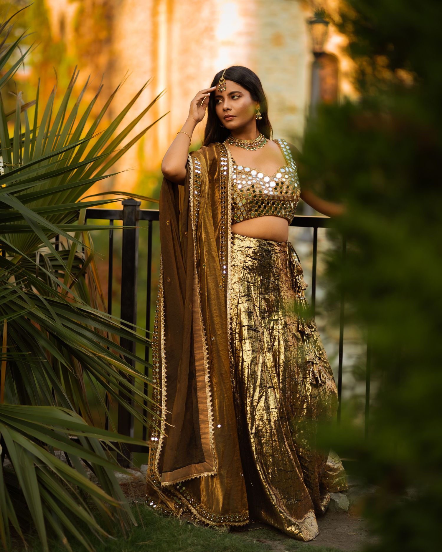 Woman in sparkly gold dress poses among palm leaves at sunset.