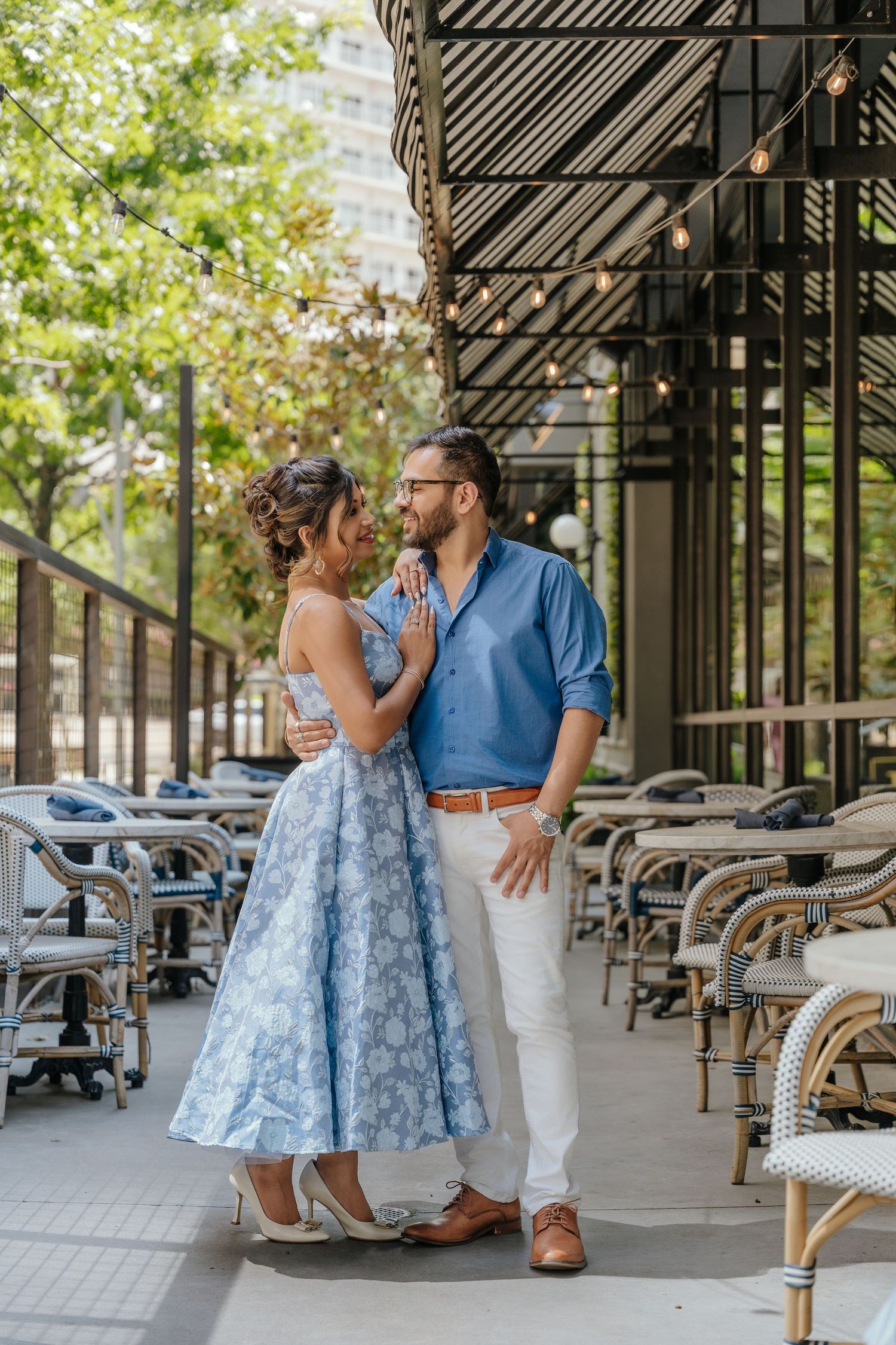 Couple in coordinating blue outfits embracing on a restaurant patio surrounded by bistro chairs and string lights.