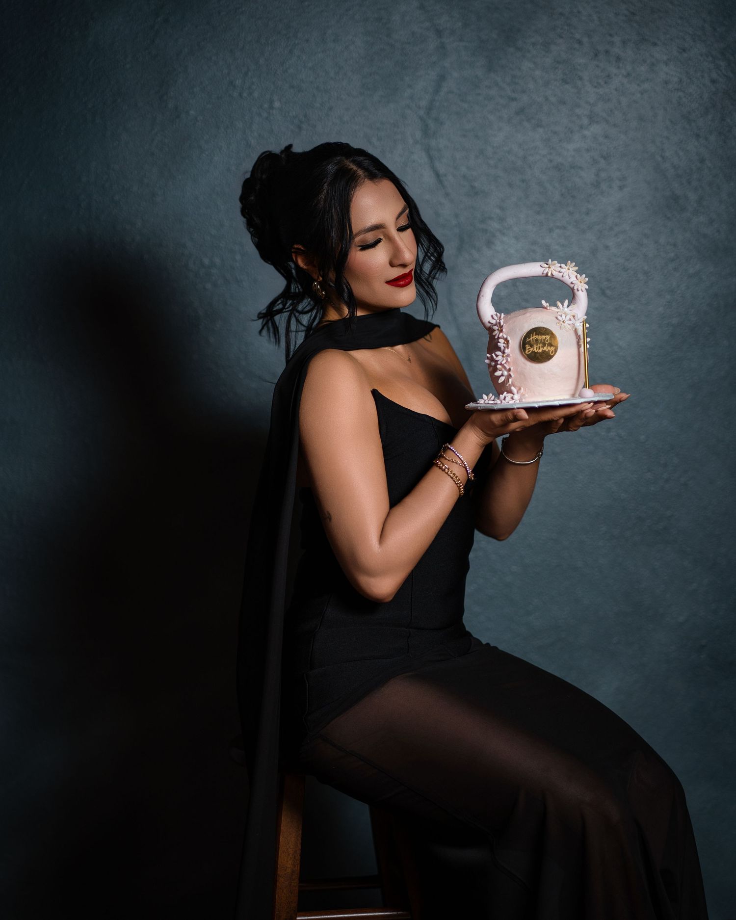 Woman in black dress sitting on stool holding decorative white birthday cake against dark blue background.